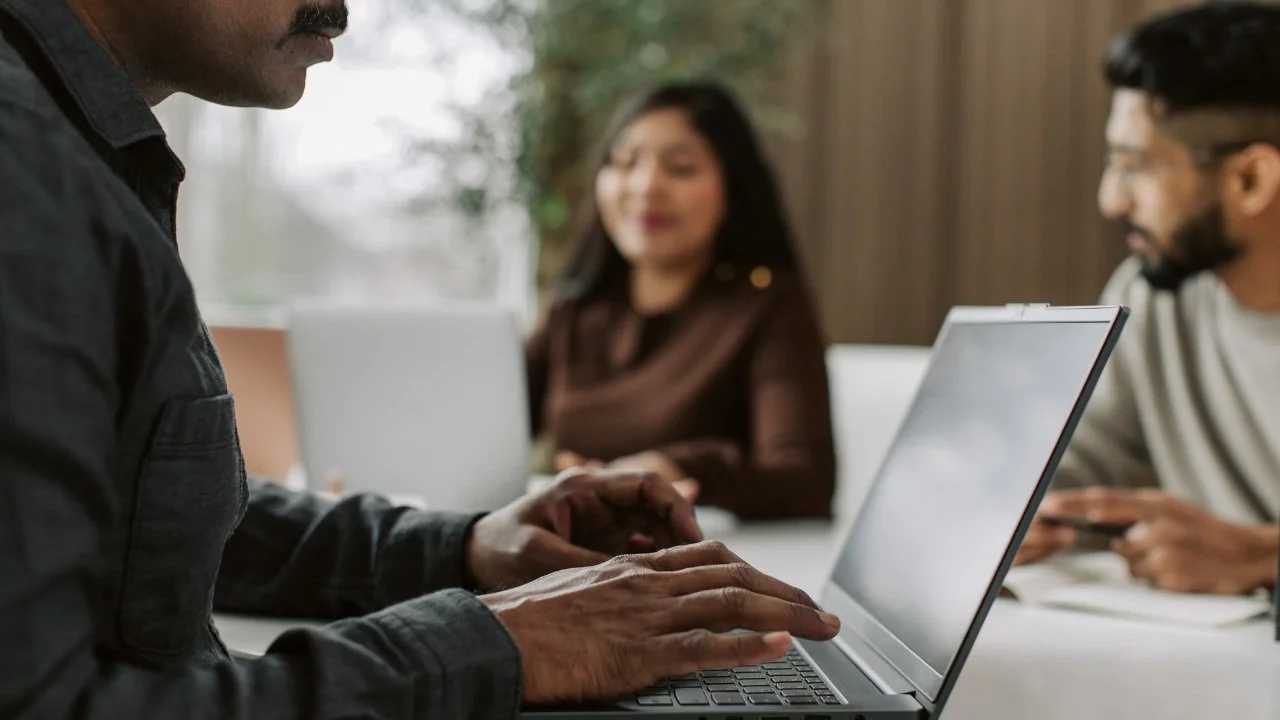 Three people in a meeting, one person typing on a laptop, another holding a phone, and a woman sitting in the background, smiling.