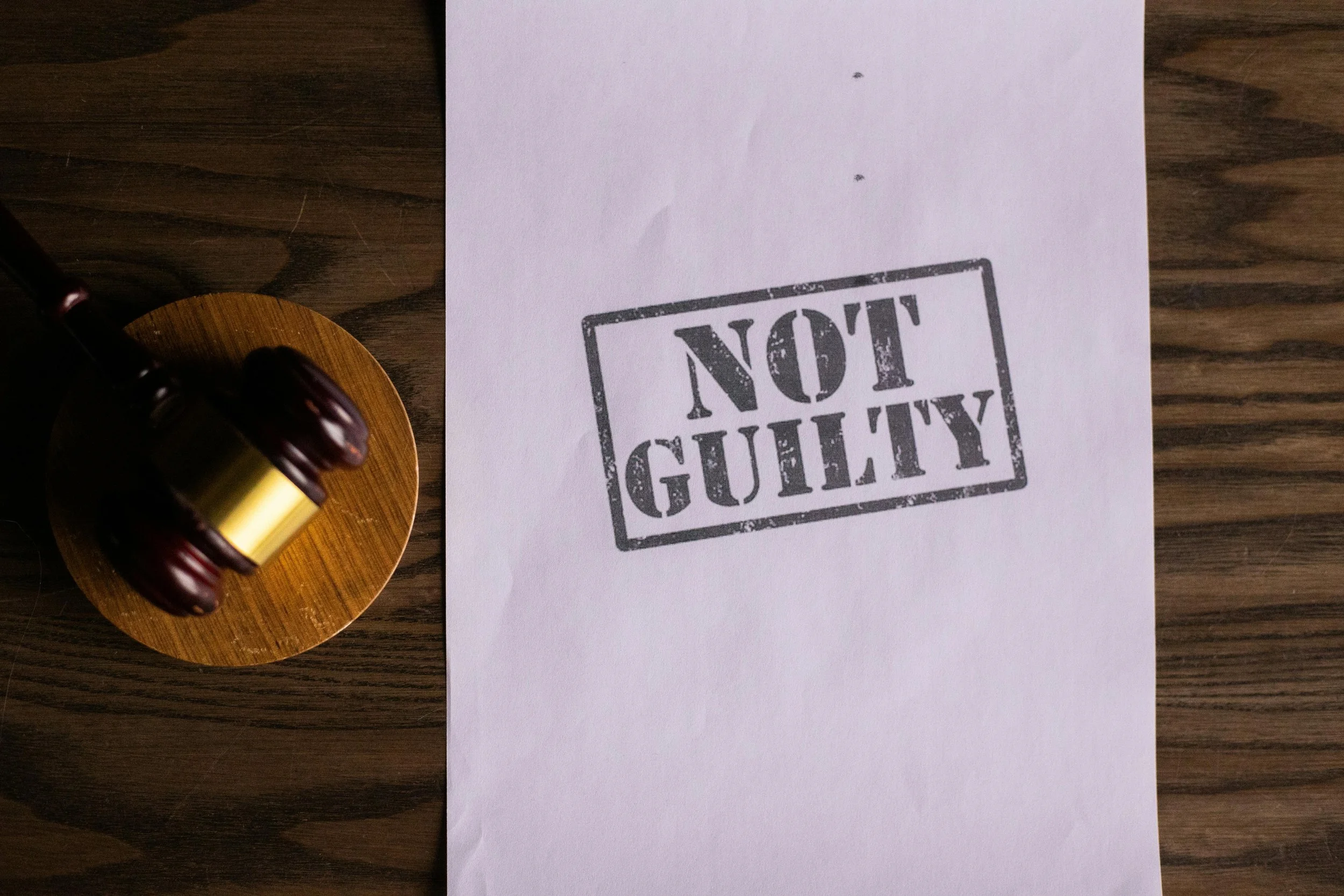 A white sheet of paper with a stamp that reads 'NOT GUILTY', placed on a wooden surface. To the left, part of a gavel and sound block are visible.