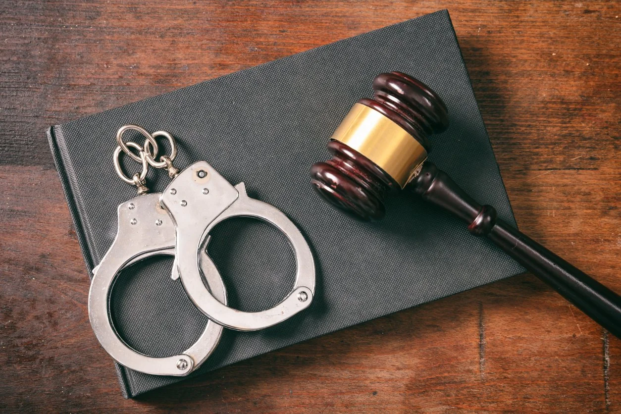 A gavel, a set of silver handcuffs, and a black book on a wooden surface.