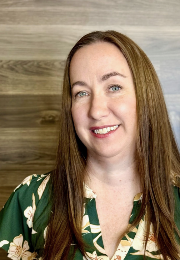 A woman with long brown hair, blue eyes, and a light complexion, smiling in front of a wooden background, wearing a green floral blouse.