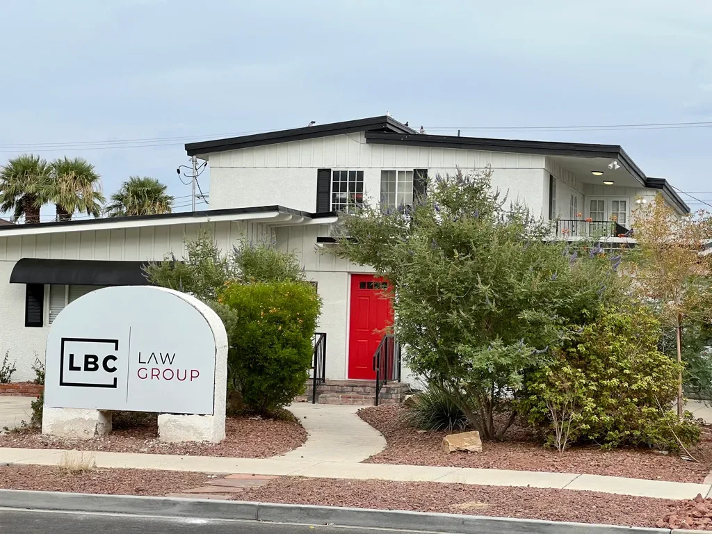 A two-story white building with a red front door and black accents, surrounded by bushes on a landscaped yard with a sign that reads 'LBC Law Group' in front.
