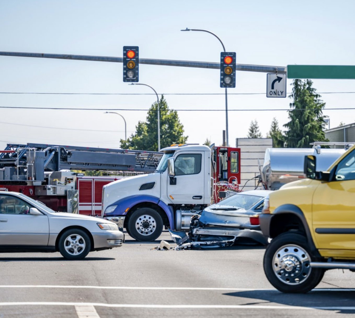 Multiple vehicles involved in a car accident at an intersection, including a damaged gray car, a large white truck, a yellow pickup truck, and a fire rescue vehicle parked nearby. Traffic lights show red signals.
