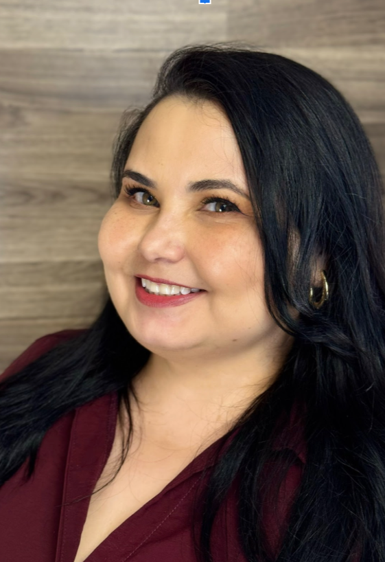 Smiling woman with long black hair wearing a maroon top and gold earrings, standing against a wooden wall background.