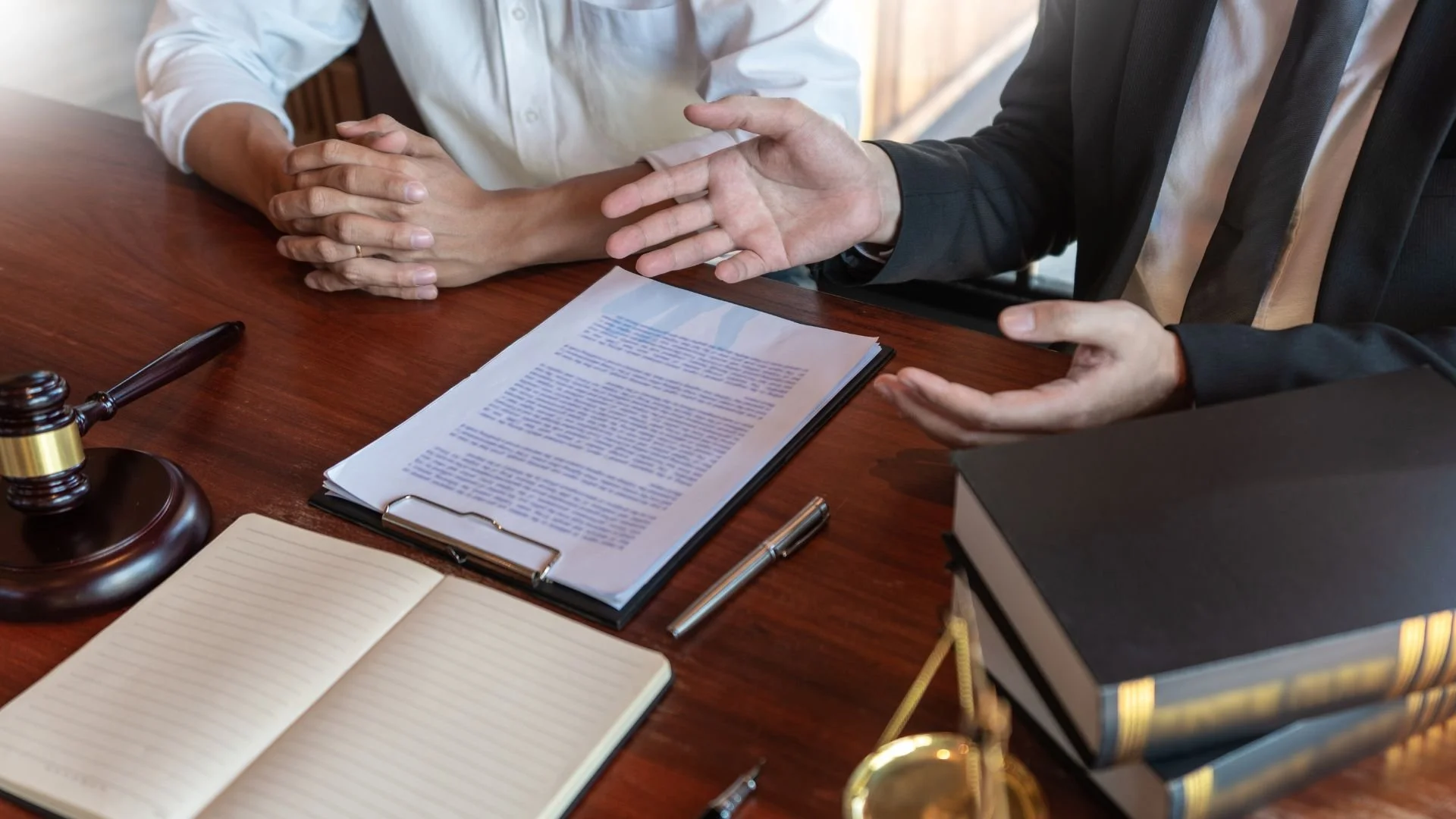 Two individuals engaged in a discussion at a wooden desk with legal documents, a gravel gavel, notebooks, and books present.