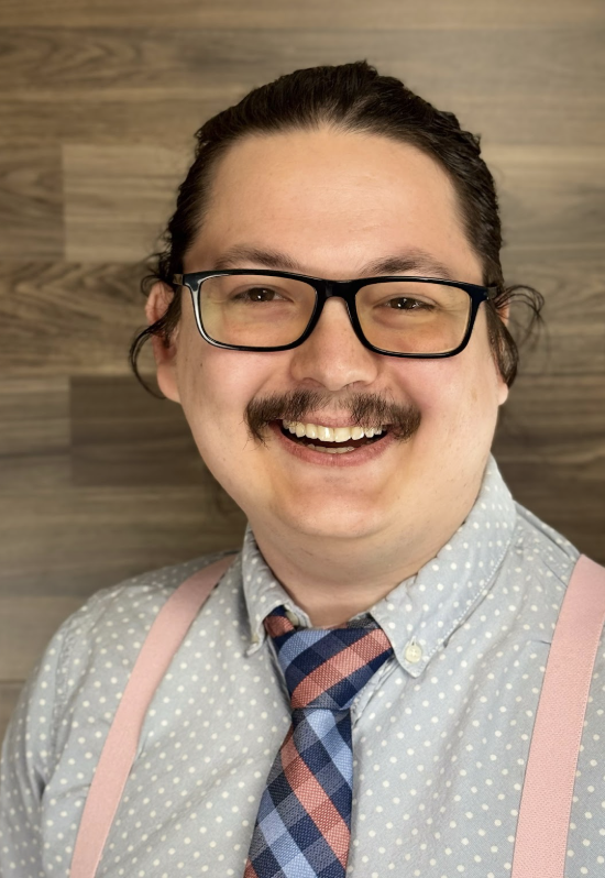 A man with glasses, a mustache, and a goatee, smiling with visible teeth. He has dark hair tied back, wearing a light blue polka dot shirt, a plaid tie, and pink suspenders. The background is a wooden wall.