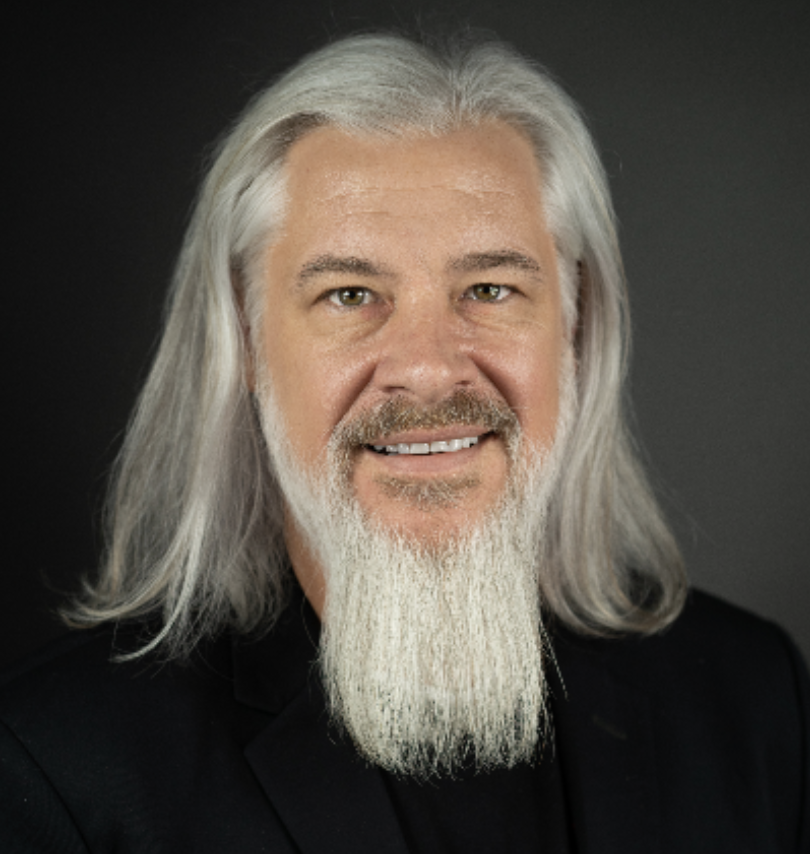 Headshot of an older man with long gray hair, a full gray beard, light-colored eyes, wearing a black shirt, against a dark background.