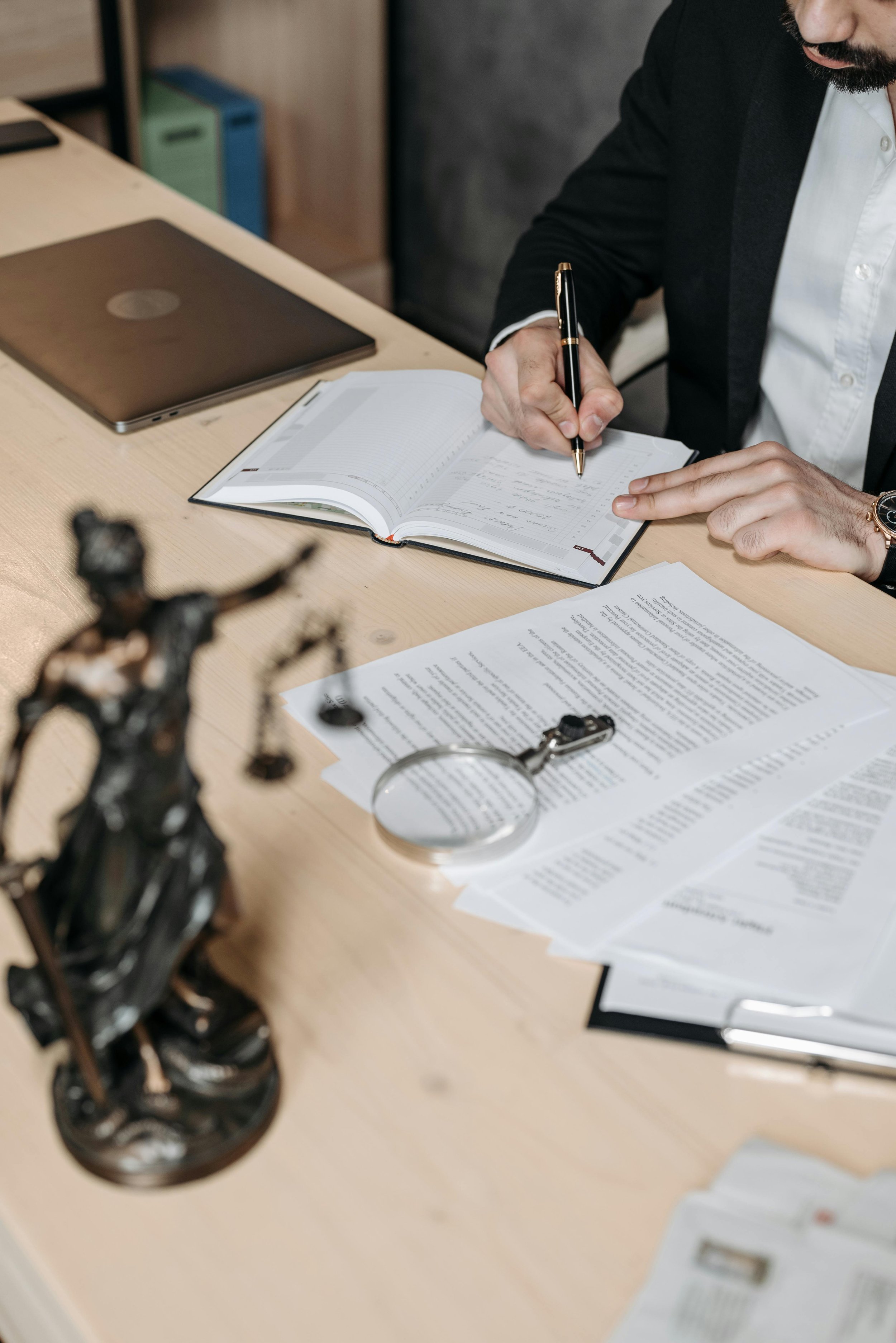 An attorney is sitting at a desk, writing in a notebook with legal documents and a magnifying glass nearby, in an office setting.