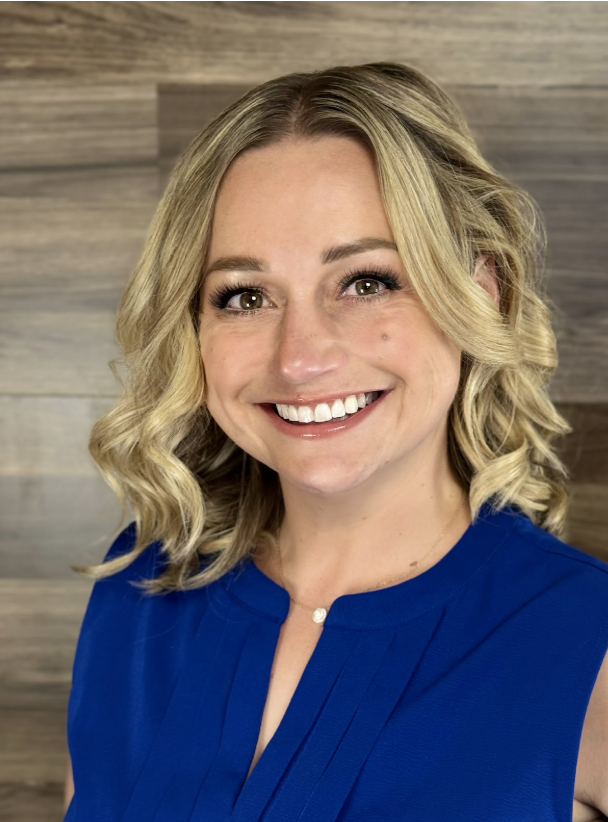 A smiling blonde woman with wavy hair wearing a blue top, standing in front of a wooden wall background.