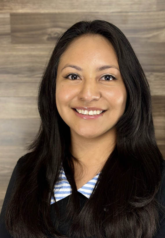 A woman with long dark hair smiling, wearing a black blazer over a blue and white striped shirt, standing in front of a wooden wall background.