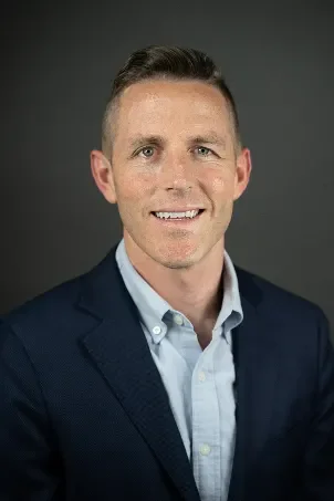 Headshot of a man wearing a dark blazer and light blue shirt, smiling against a dark gray background.