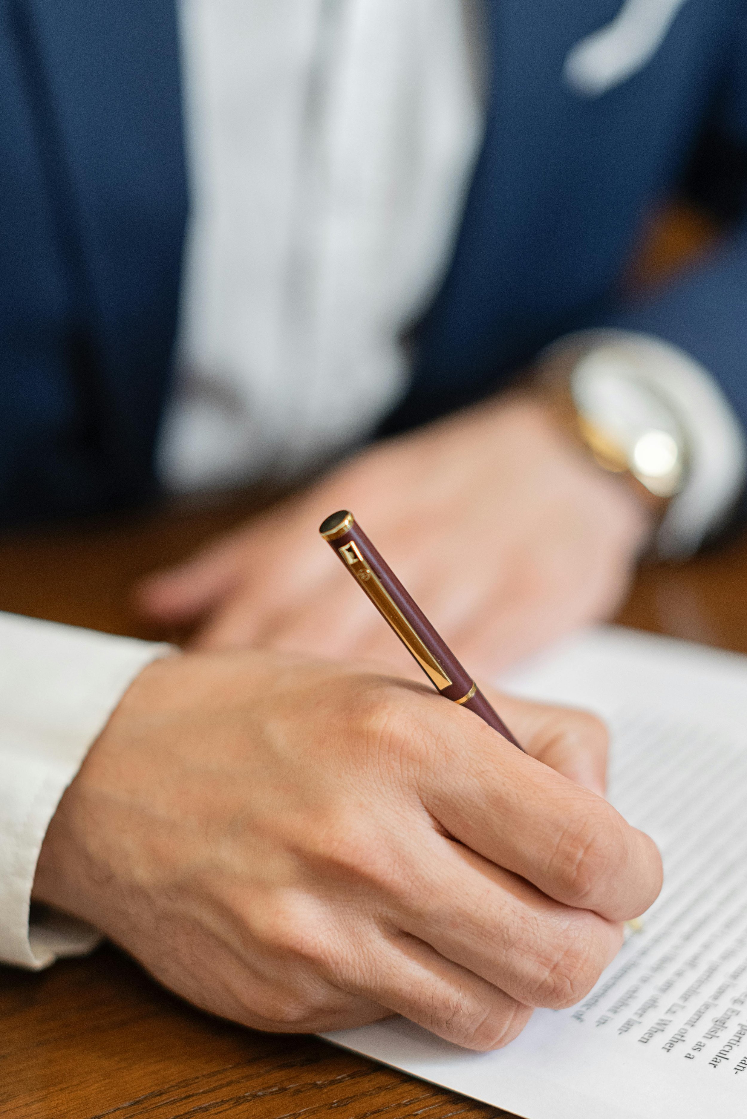 Close-up of a person wearing a suit and watch, signing a document with a pen on a wooden table.