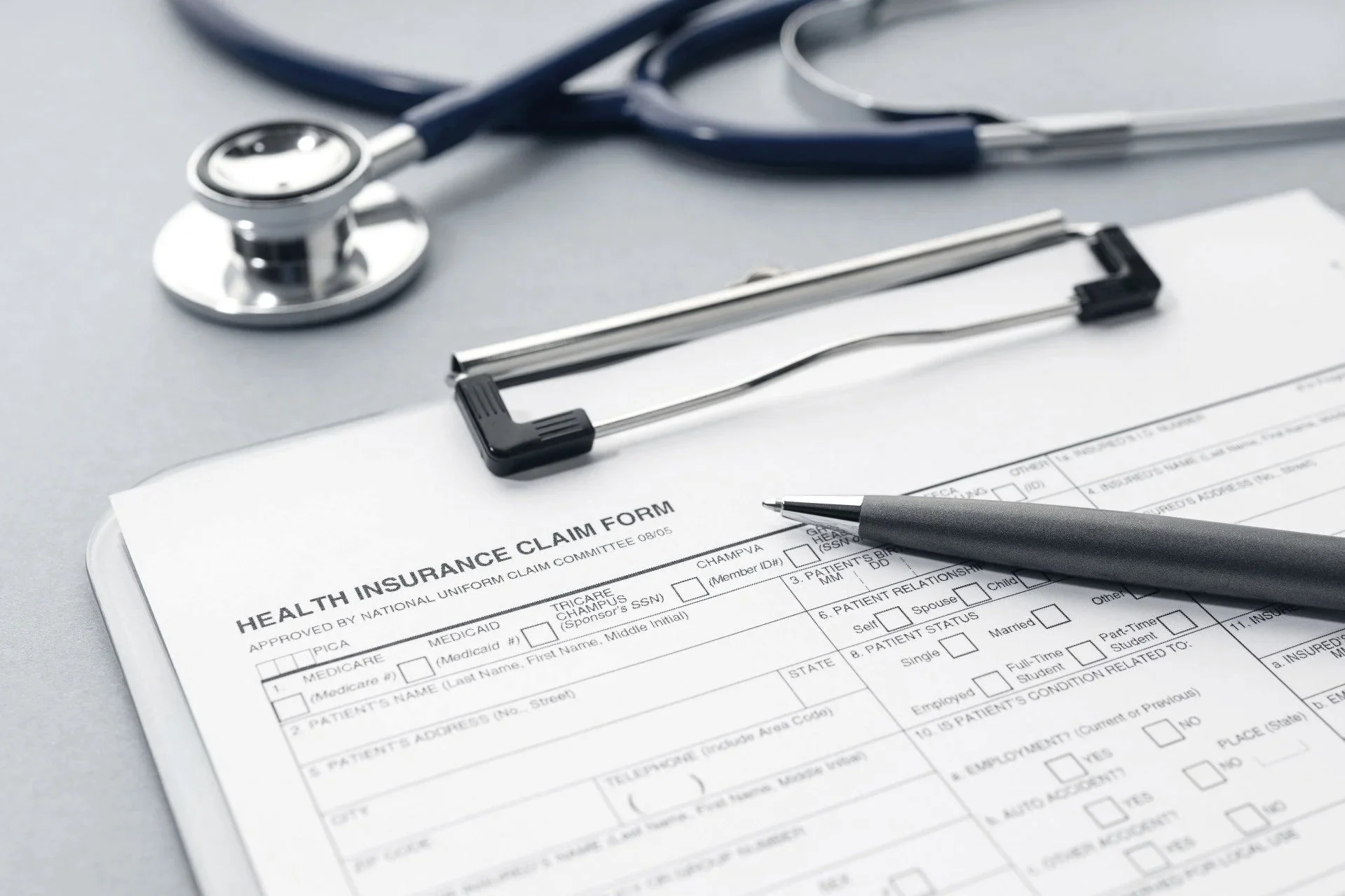 A doctor's stethoscope resting on a desk next to a clipboard holding a health insurance claim form and a pen.