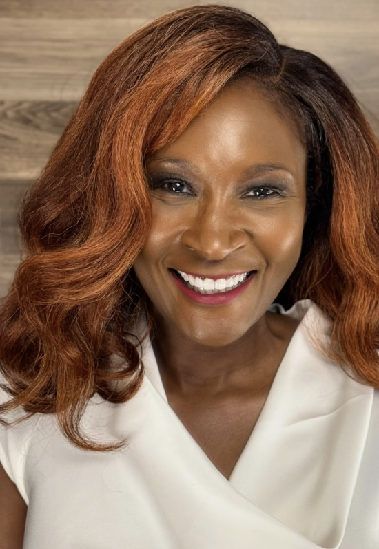 Close-up of a smiling African American woman with long, wavy reddish-brown hair, wearing a white top, with a wooden background.