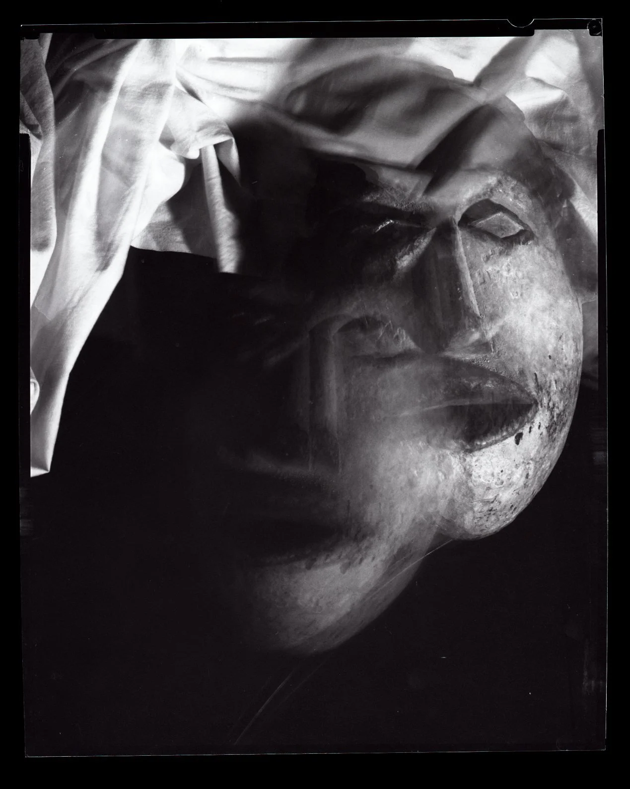 A close-up black and white photograph of a carved wooden mask of a human, partially covered with a cloth material.