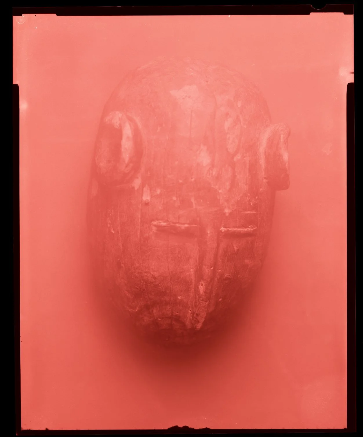 An ancient carved wooden mask with features resembling a human face, displayed on a pink background