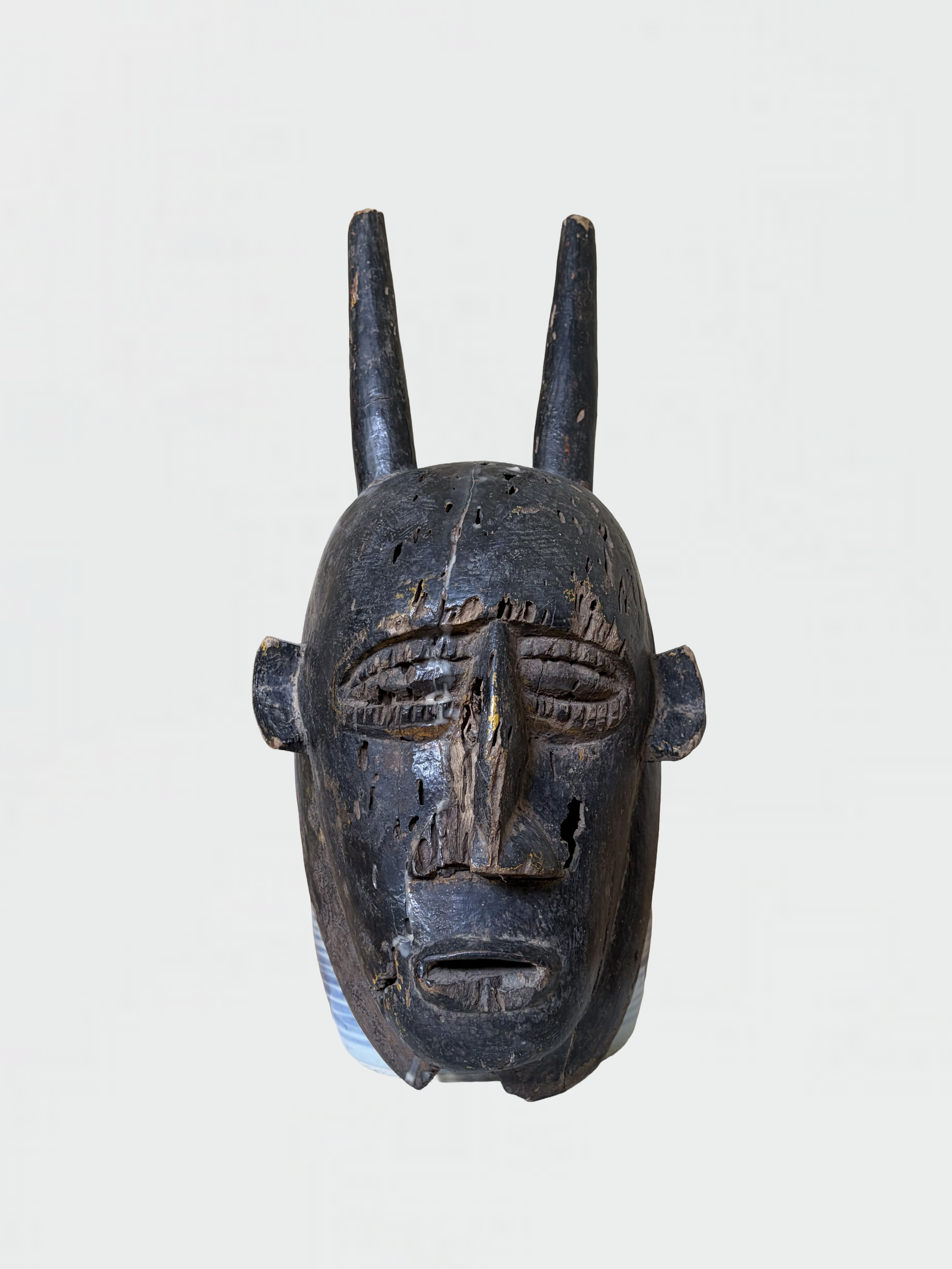 An ancient wooden African tribal mask with carved facial features, painted black with some worn areas, and featuring two tall vertical horns on top.