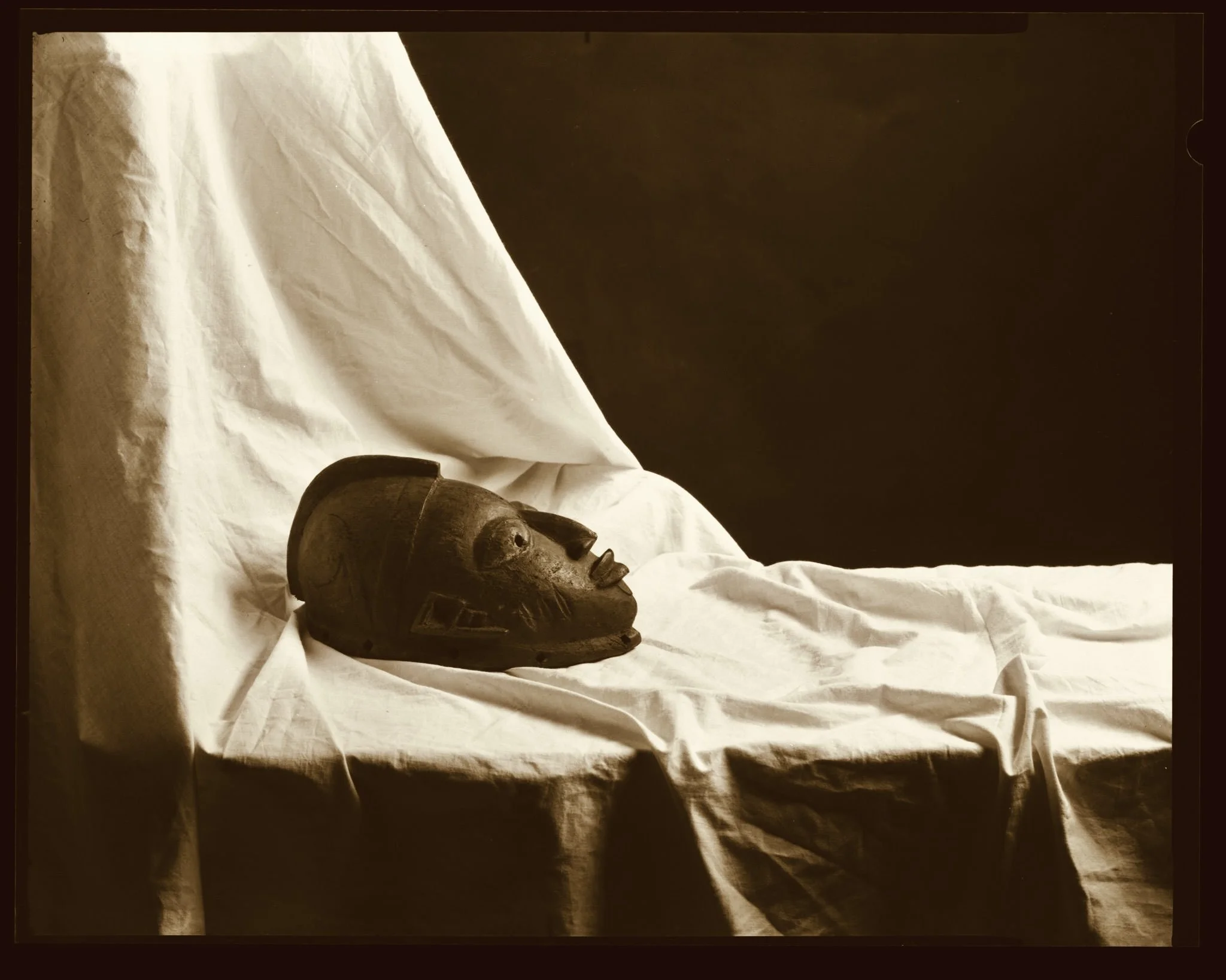 A carved wooden African mask resting on a white cloth, with a dark background.