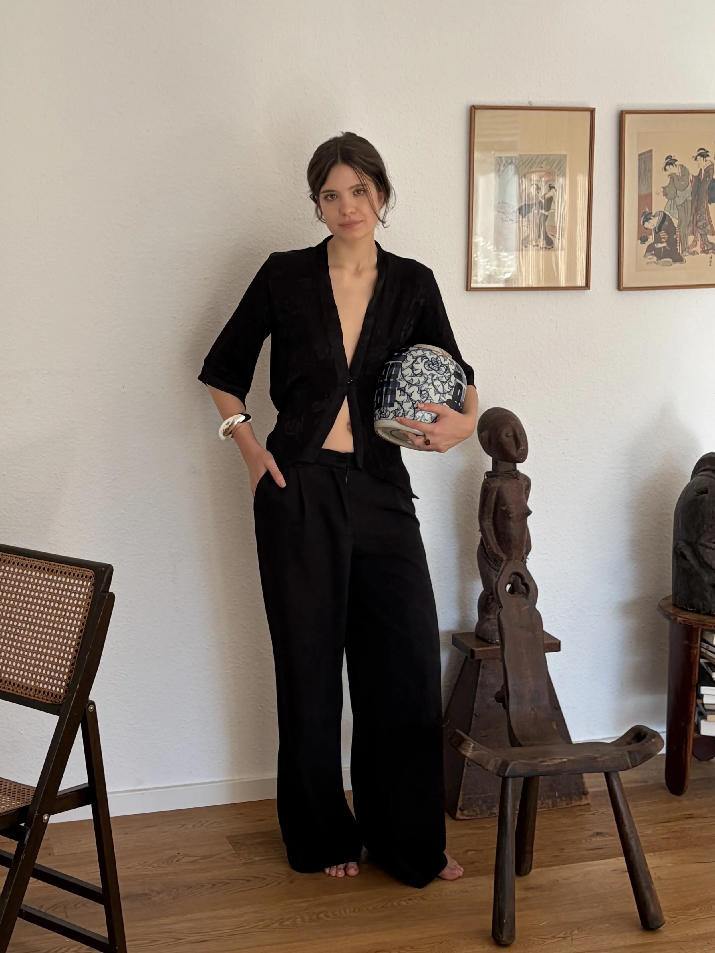 A woman with short dark hair wearing a black top and black pants holding a patterned bowl stands barefoot next to a wooden carved sculpture and a wooden chair, in a room with white walls and framed artwork.