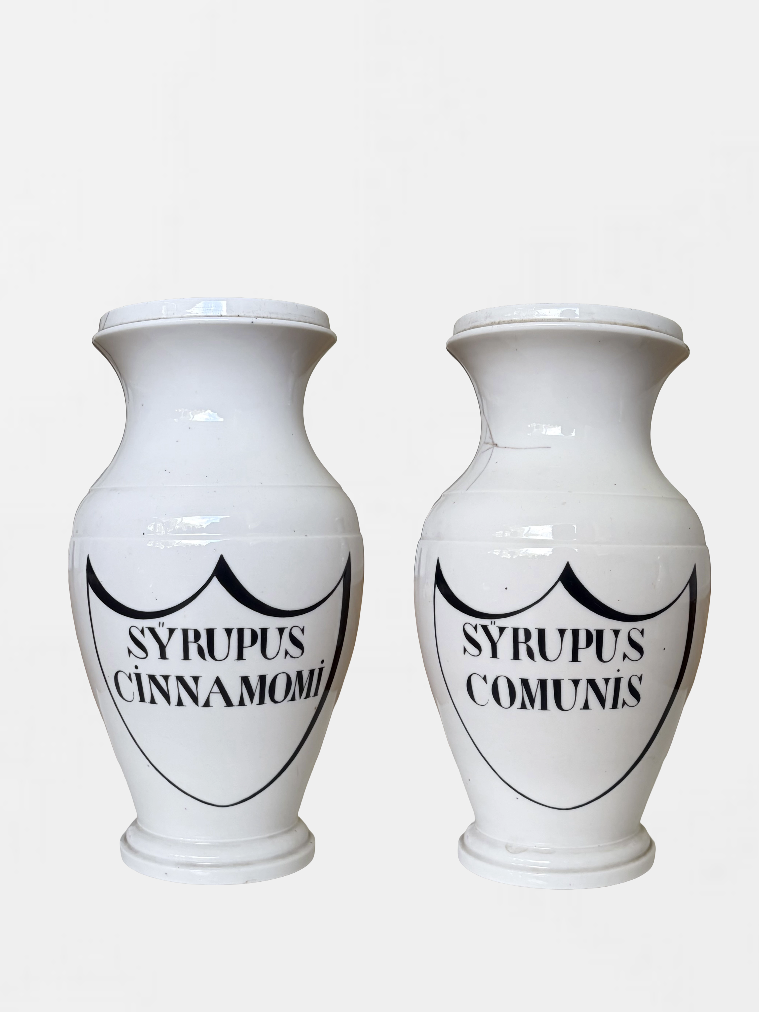 Two white ceramic jars with black decorative lettering and designs, one labeled "SYRUPUS CİNNAMOMI" and the other labeled "SYRUPUS COMUNIS" on a plain white background.