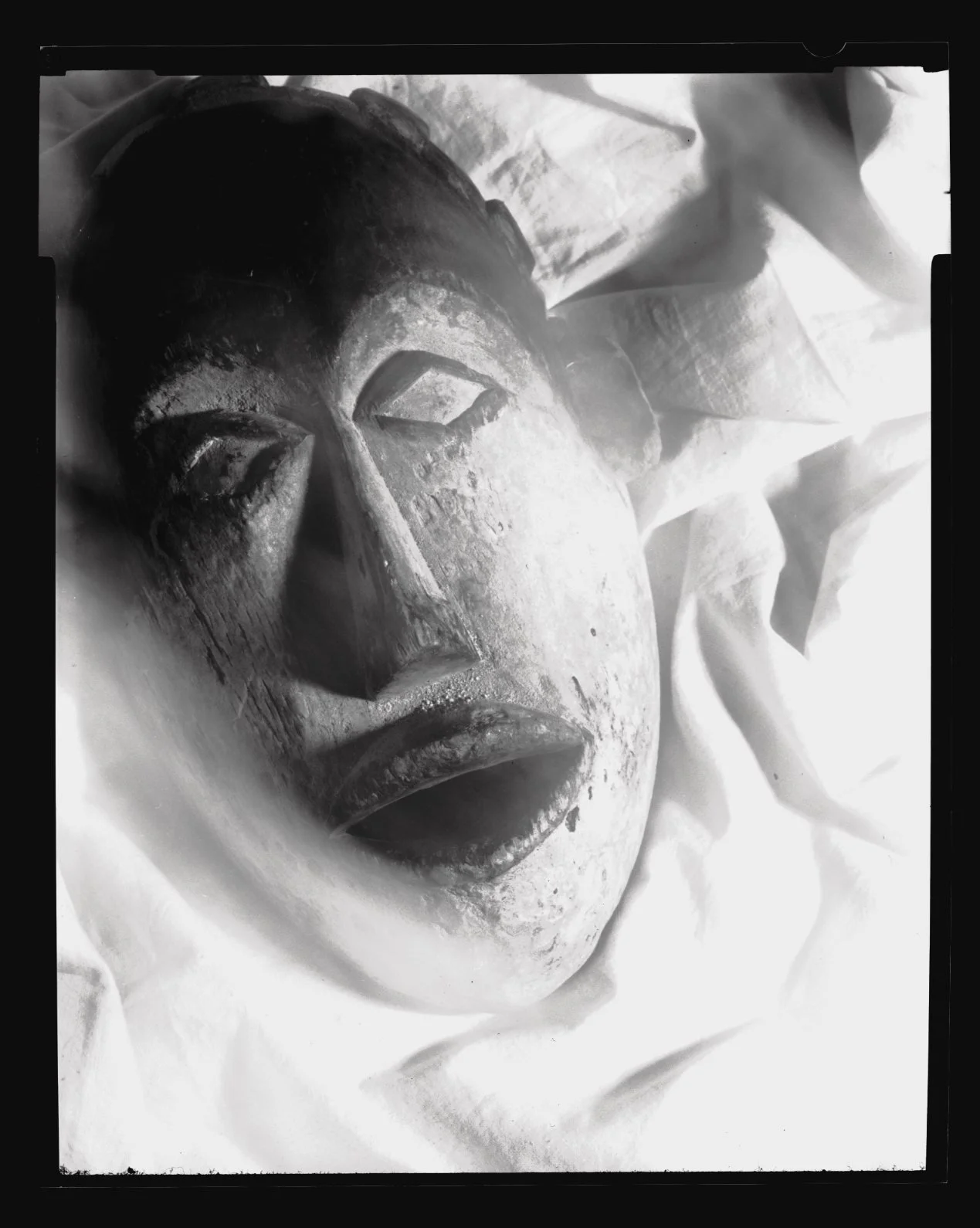 Black and white photograph of an Africanmask with closed eyes and detailed facial features, resting on a white cloth.