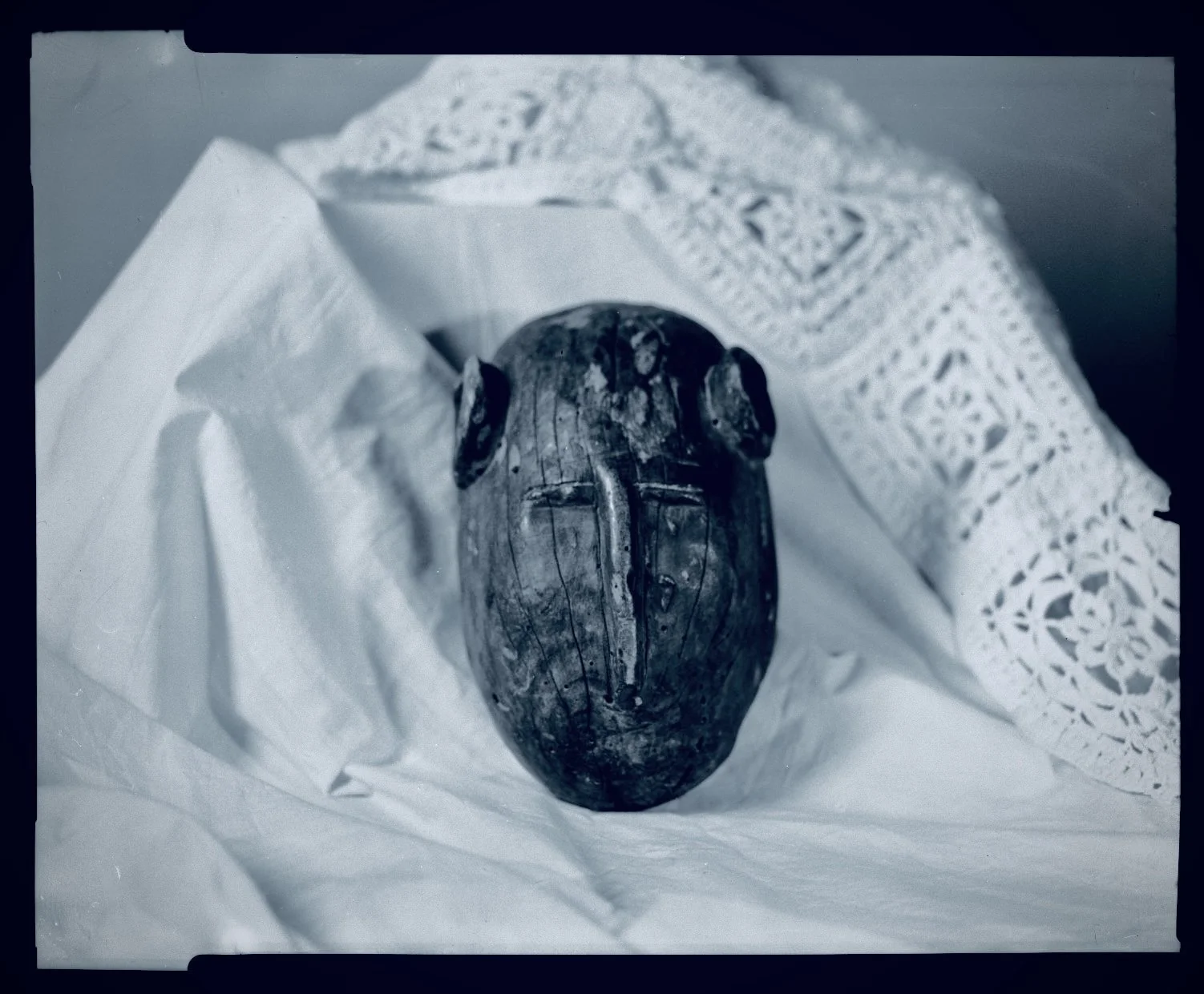 A wooden carved mask with elongated face, closed eyes, and a prominent nose, placed on white paper and decorative fabric.