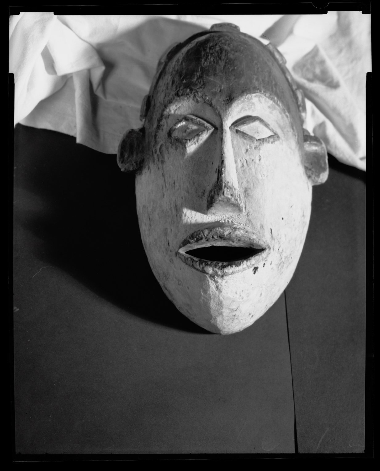 Black and white photo of a carved wooden tribal mask with a wide open mouth, closed eyes, and large ears, placed on a flat surface with crumpled white cloth in the background.