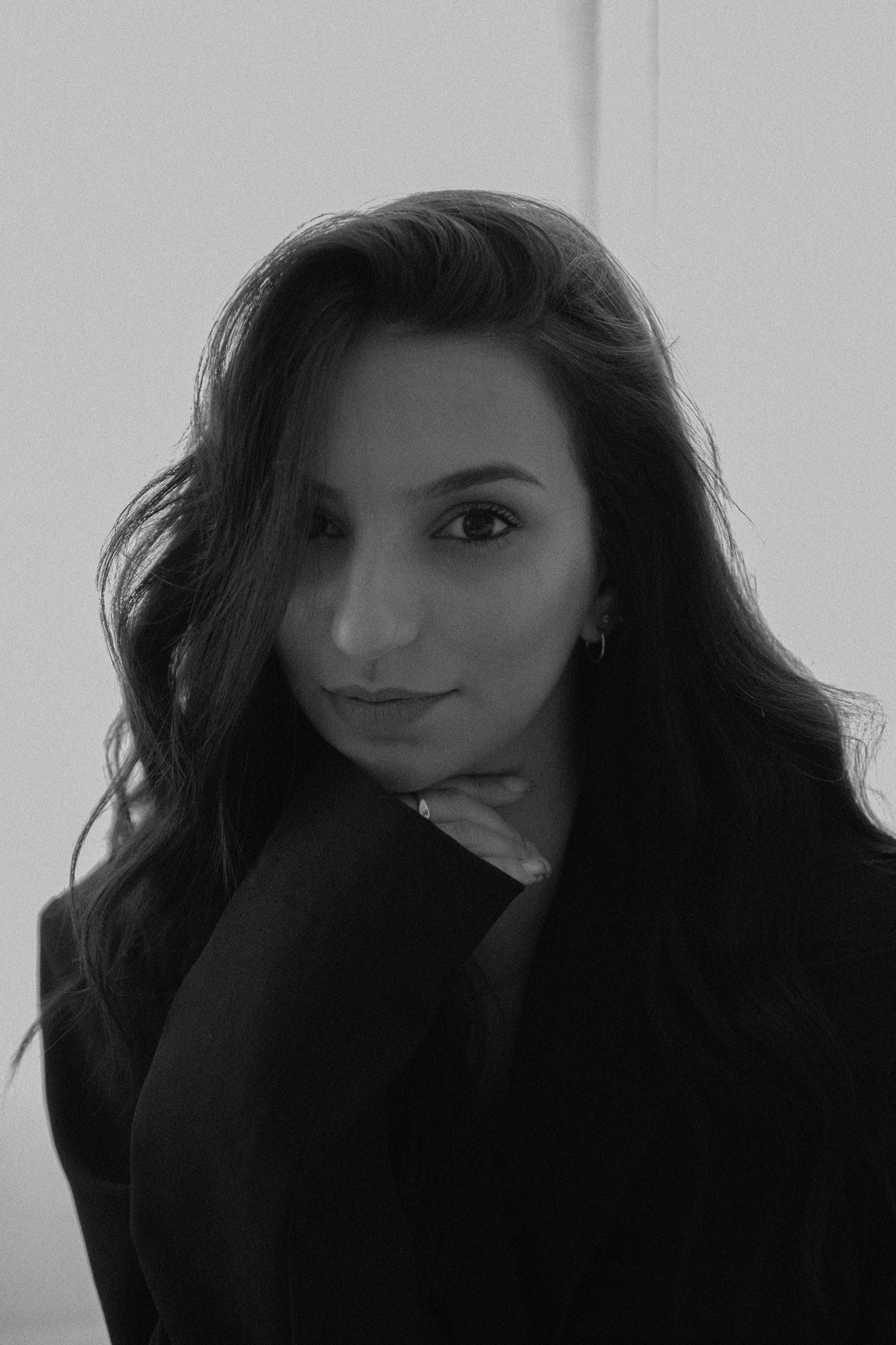 Portrait d'une femme aux cheveux longs et ondulés, portant un blazer noir, souriant légèrement avec la main sous le menton, en noir et blanc.