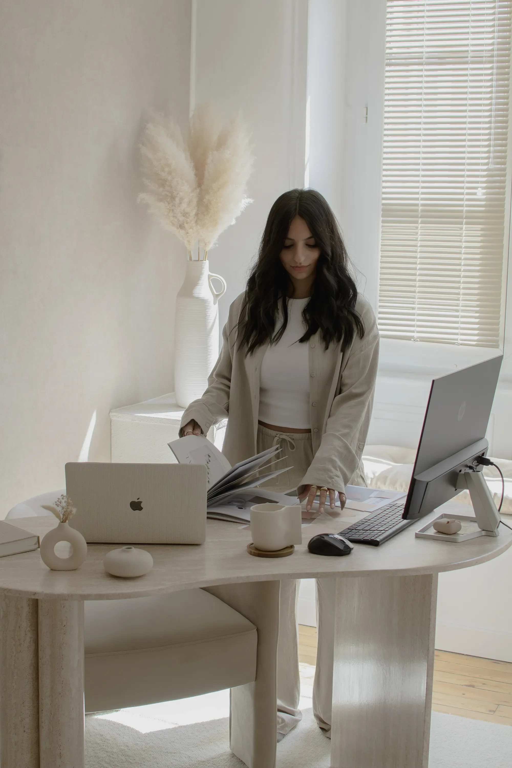 Une femme avec des cheveux foncés et longs travaille à un bureau beige avec un ordinateur portable, une tablette, des cahiers et des objets décoratifs beige dans une pièce lumineuse avec des volets beige