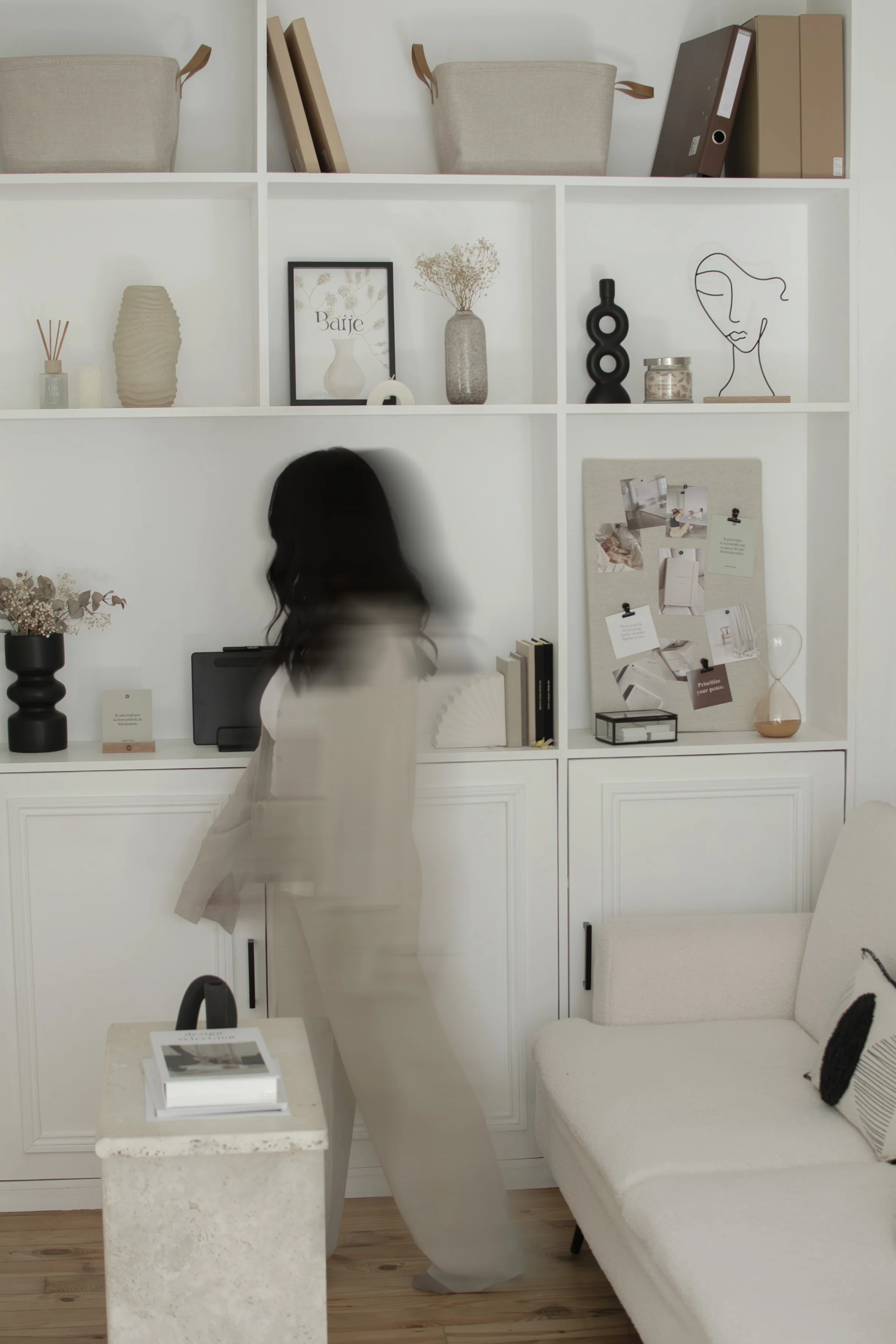 Une femme se tenant dans un salon minimaliste avec une bibliothèque blanche remplie de décorations comme des vases, des dessins et des livres, et un canapé blanc.