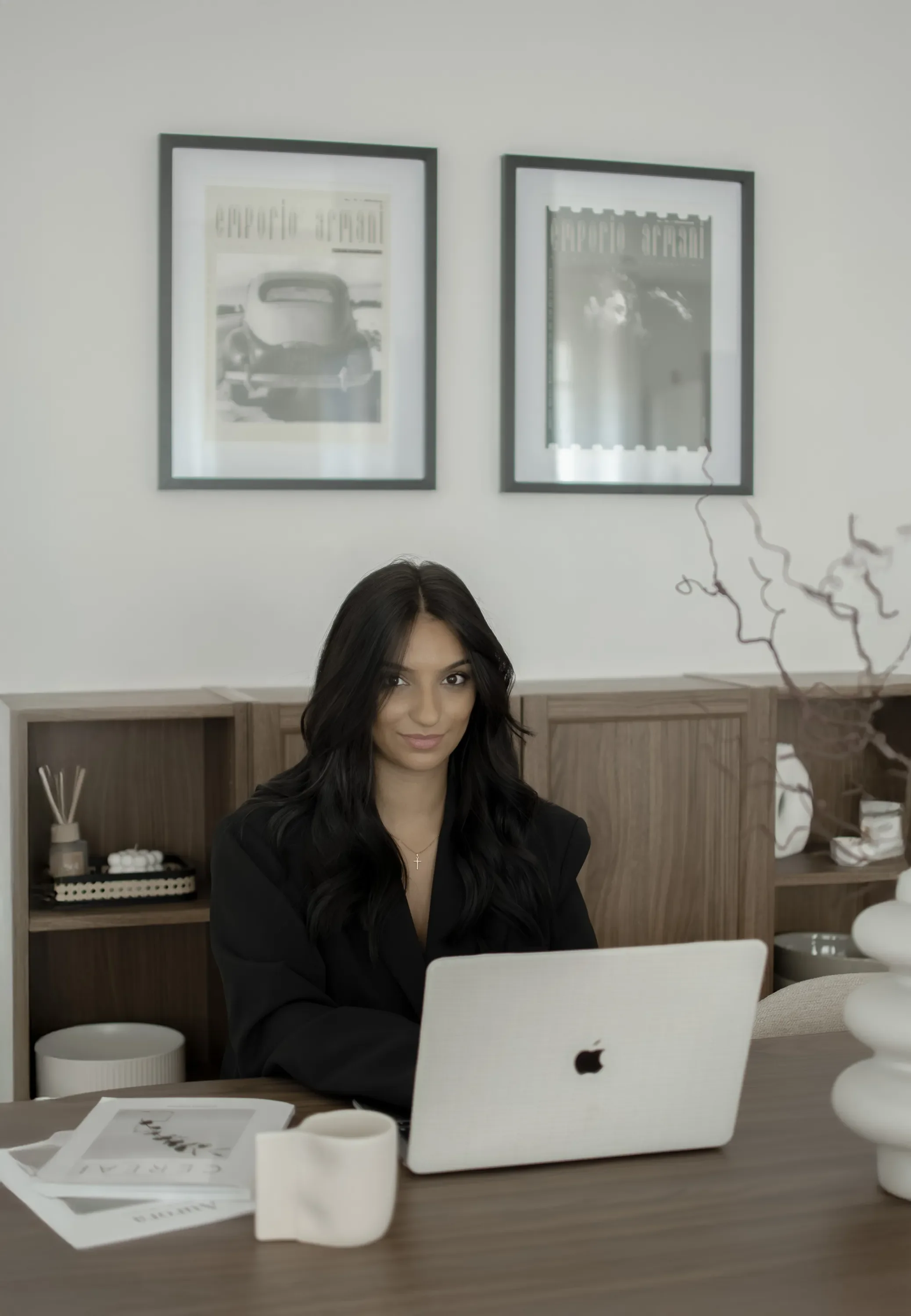 Une femme aux cheveux longs et foncés, portant une veste noire, assise devant un ordinateur portable blanc Apple sur une table en bois, dans un bureau moderne avec étagères en bois clair, décor minimaliste, et deux affiches en noir et blanc encadrées au mur.