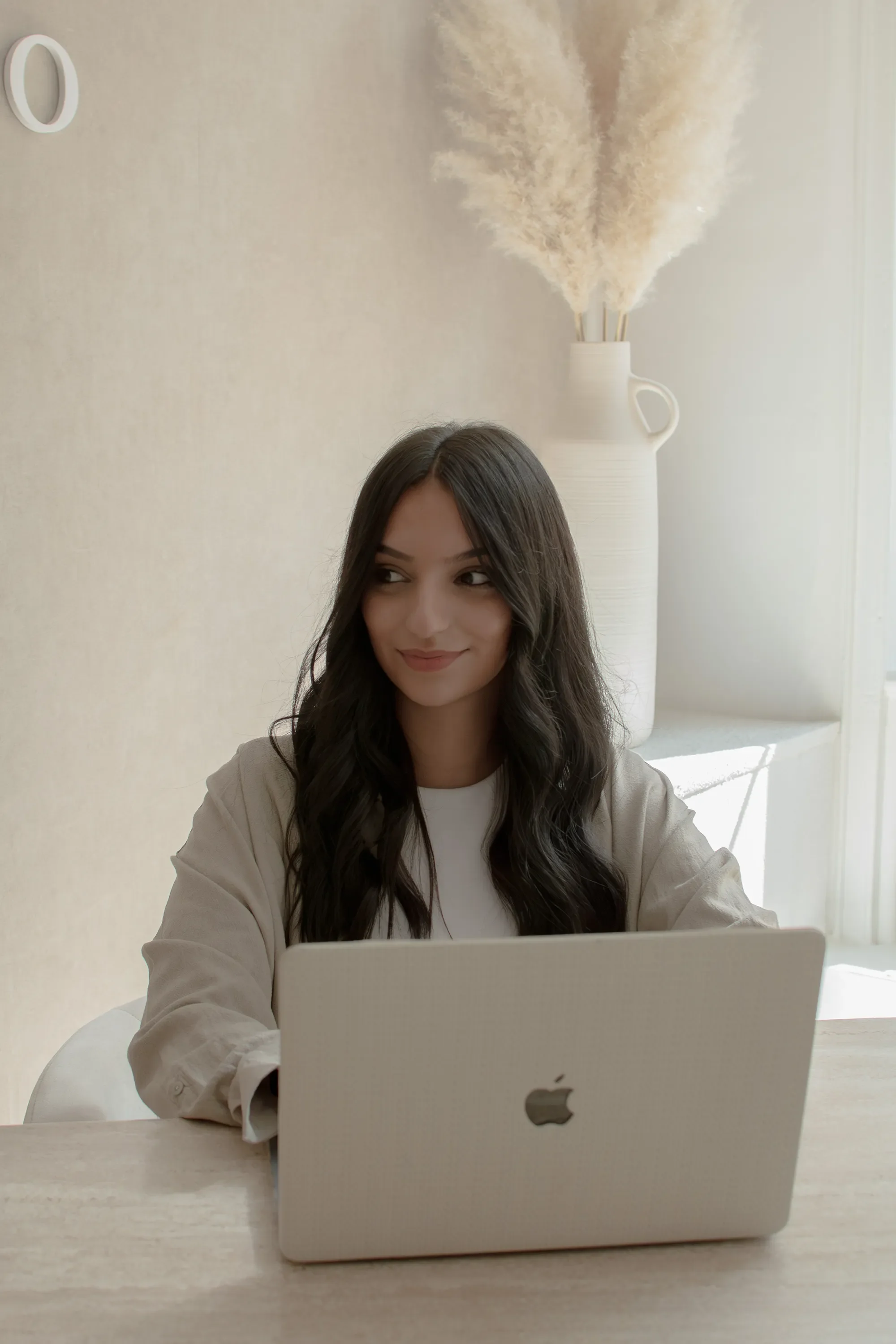 Une femme assise à une table utilisant un ordinateur portable Mac, avec un vase blanc contenant des branches de pampa en arrière-plan