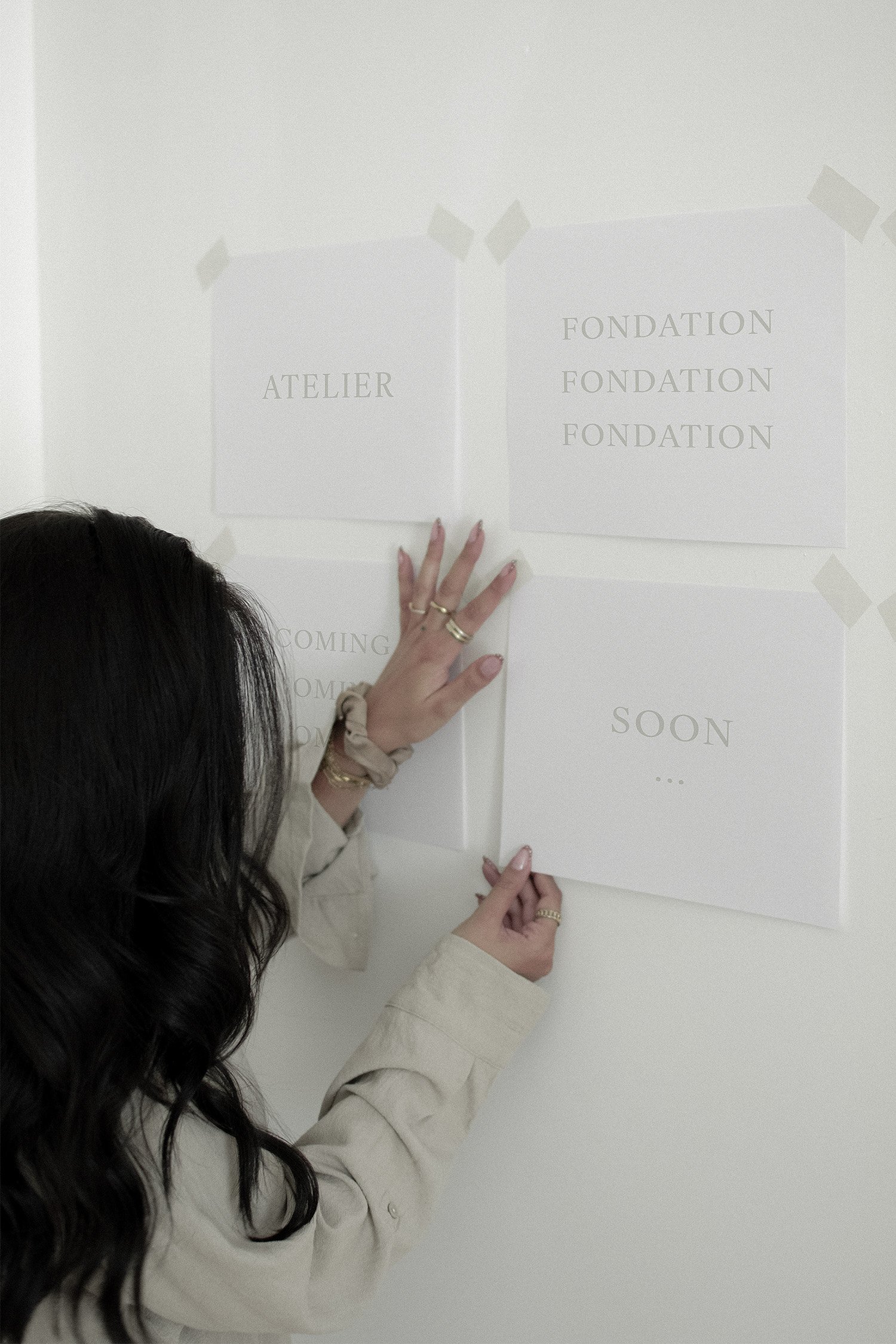 Une personne tient des affiches blanches collées au mur avec du ruban adhésif. L'une des affiches affiche le mot 'ATELIER' et l'autre affiche le mot 'FONDATION' répété plusieurs fois.