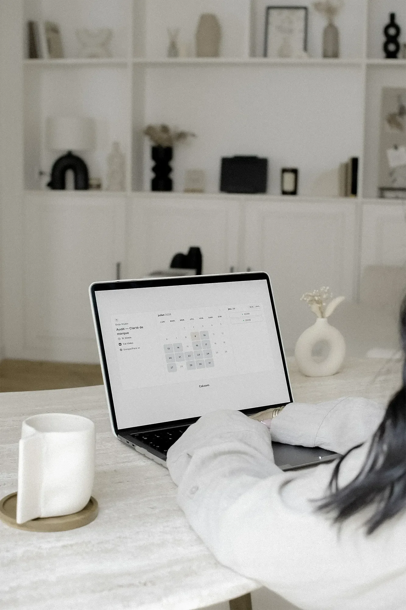 Un ordinateur portable sur une table avec un calendrier affiché à l'écran, un vase avec des fleurs et une tasse blanche. En arrière-plan, une étagère blanche avec des décorations et des objets noirs ou blancs.