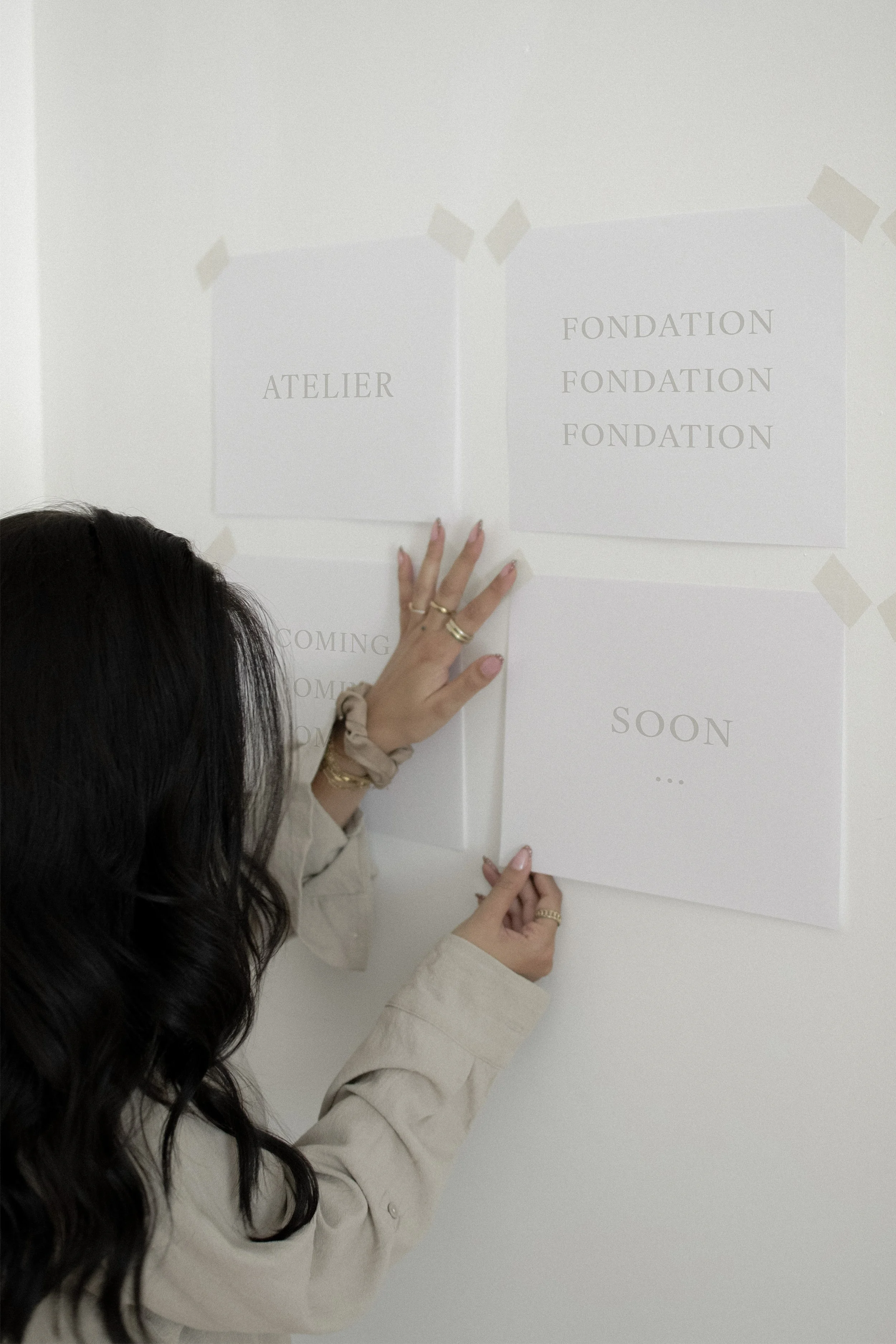 Une personne avec de longs cheveux noirs qui accroche sur un mur blanc des feuilles de papier, avec des mots 'ATELIER', 'FONDATION', "COMING" et 'SOON'