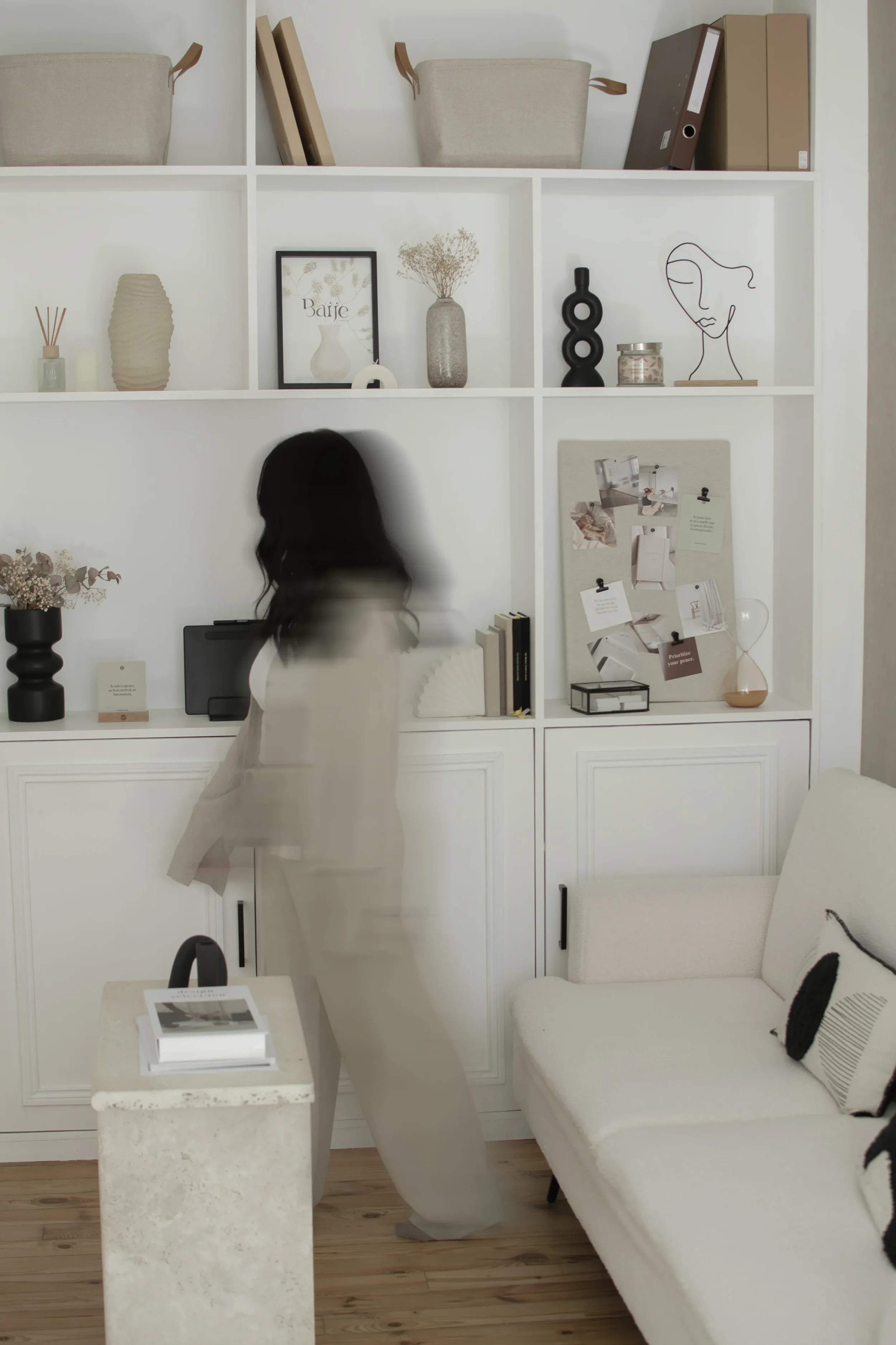 Une étagère blanche décorée avec des objets tels que des vases, des cadres, des livres, une sculpture abstraite et un tableau. En dessous, une personne avec des cheveux noirs. À droite, un canapé blanc avec un coussin noir et blanc. Au premier plan, une table en pierre avec un livre dessus.