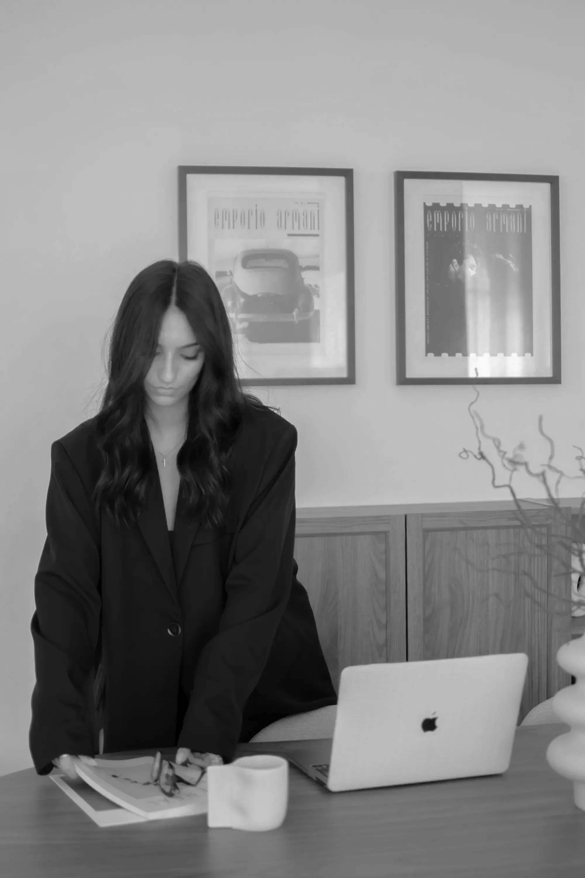 Une femme aux cheveux longs debout devant une table et un ordinateur portable Apple, avec une tasse, des lunettes, des documents, et une plante. Deux cadres sont accrochés au mur derrière elle