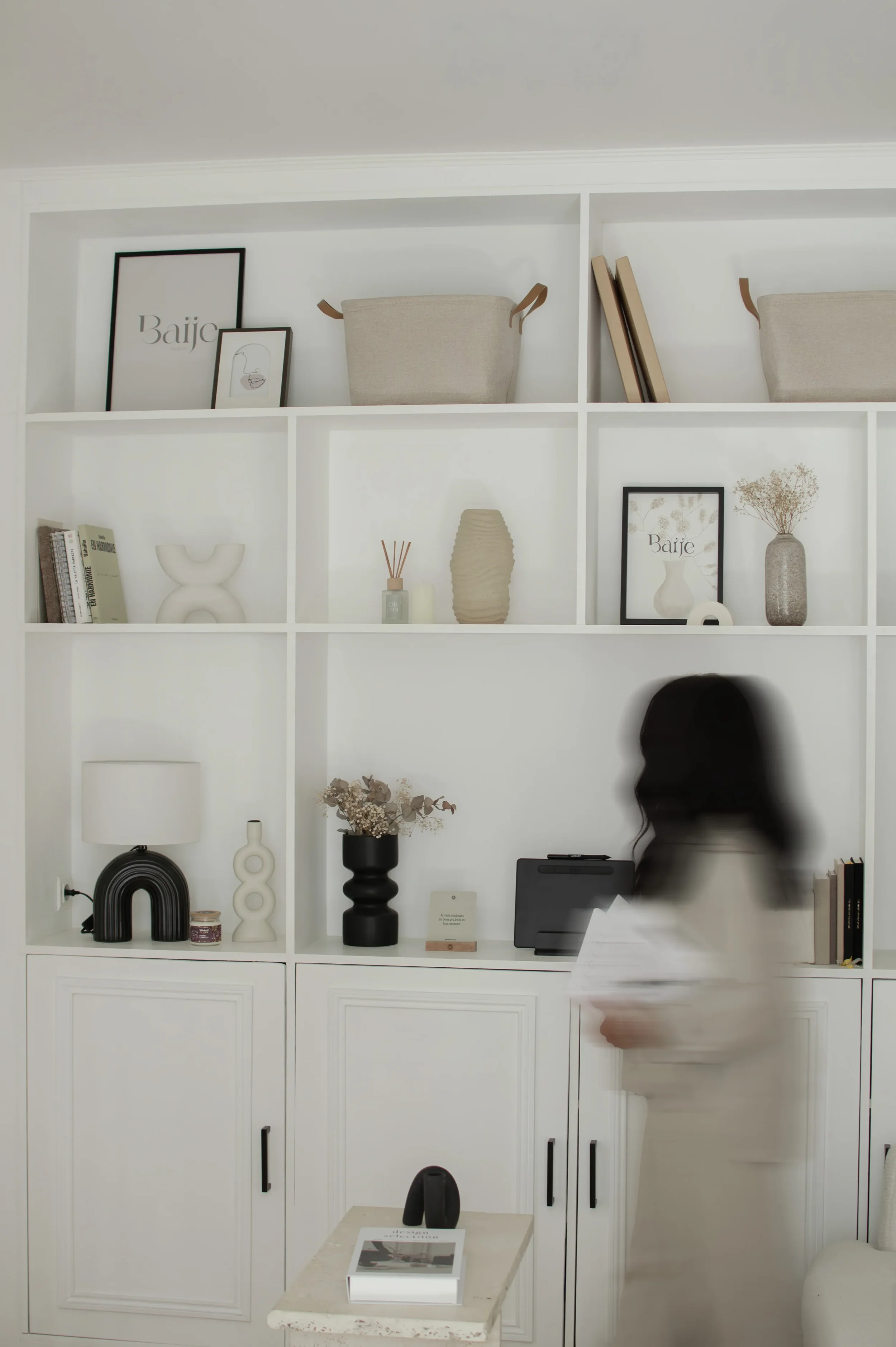 Une étagère blanche décorée avec des objets tels que des vases, des cadres, des livres, une sculpture abstraite et un tableau. En dessous, une personne avec des cheveux noirs. À droite, un canapé blanc avec un coussin noir et blanc. Au premier plan,