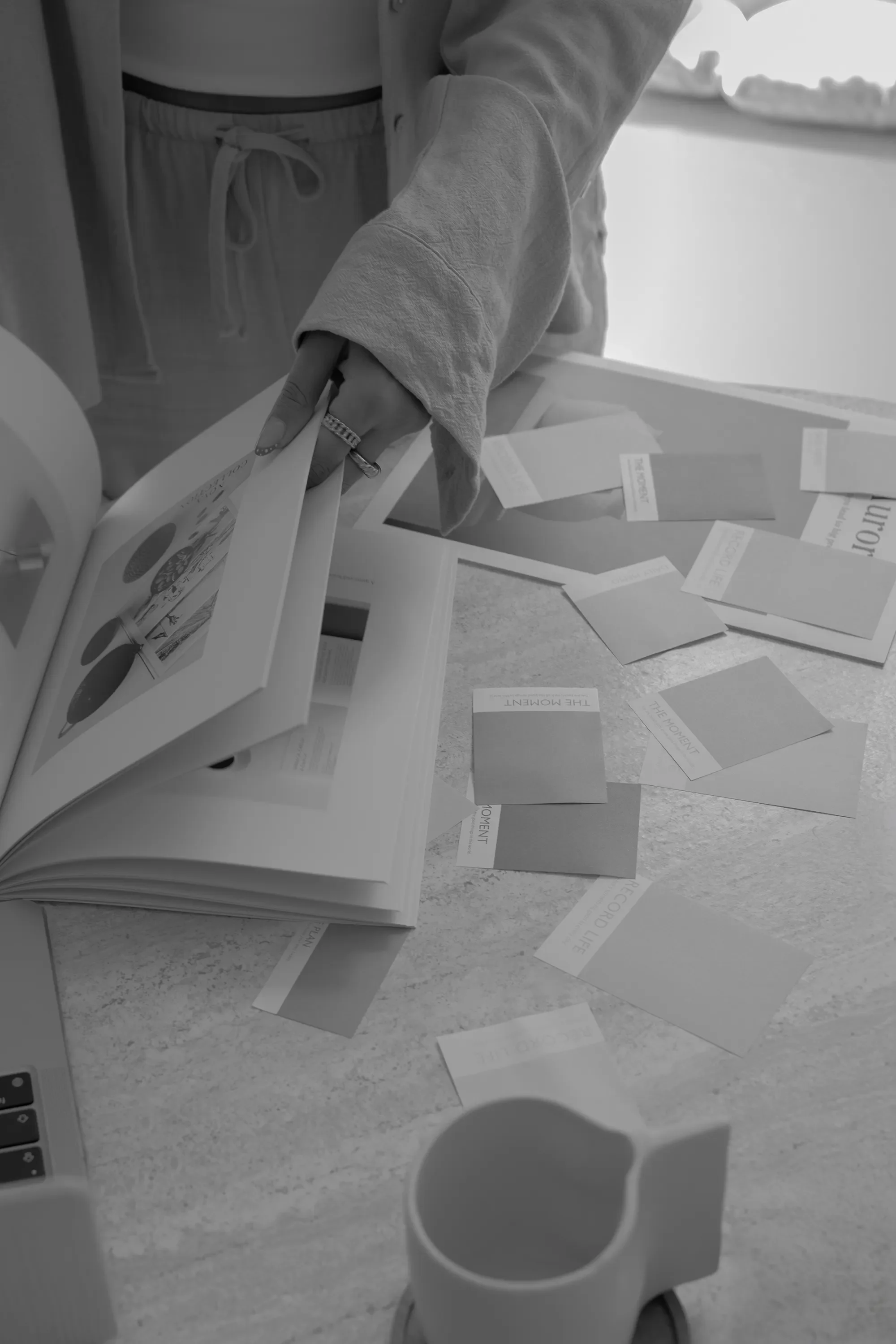 Une personne montre un livre ouvert avec des illustrations, entourée de plusieurs cartes ou papiers dispersés sur une table, vue partielle en noir et blanc.