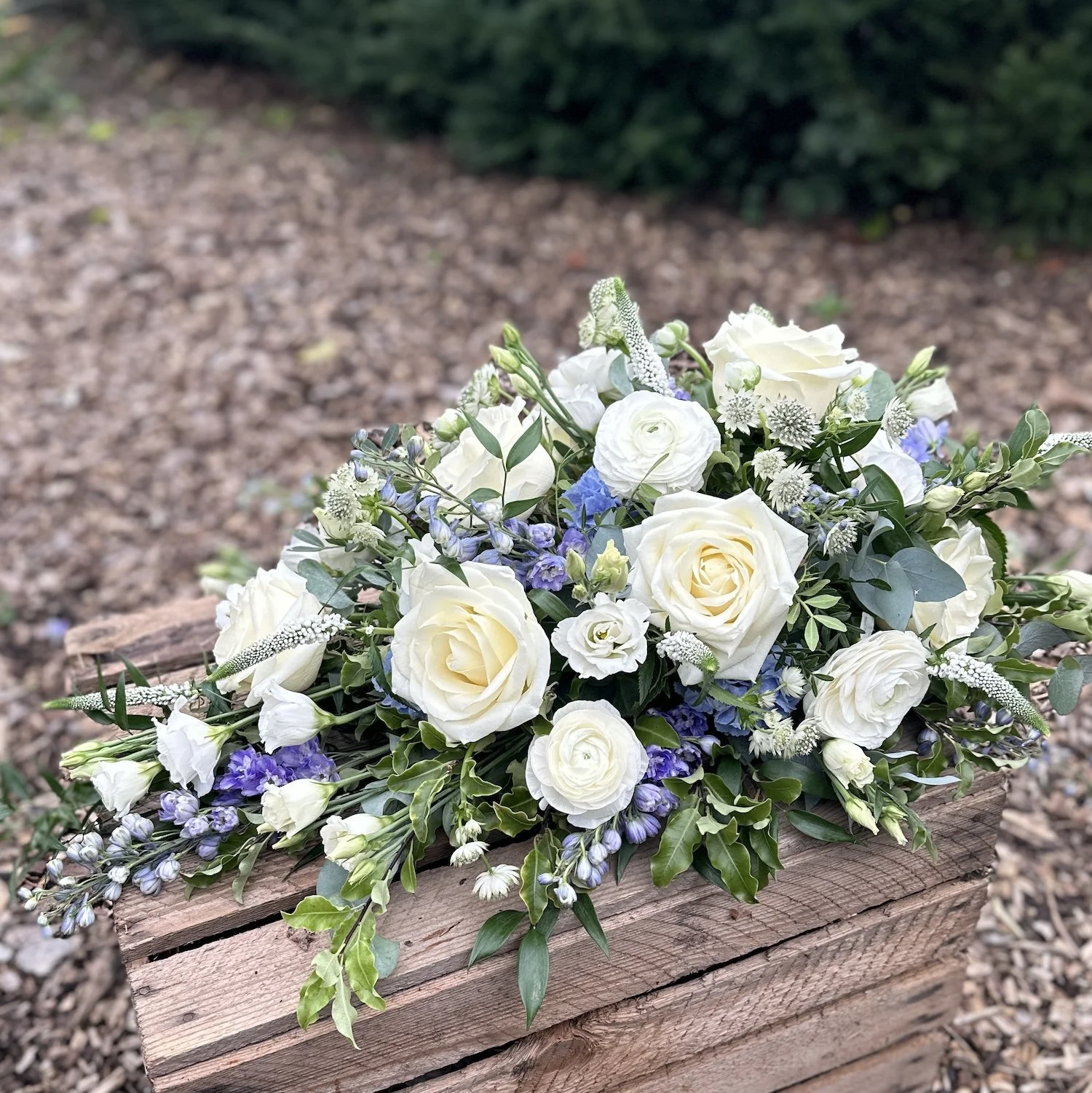 A small casket spray of white roses, purple, and white flowers with green foliage in a wooden crate on a gravel surface.