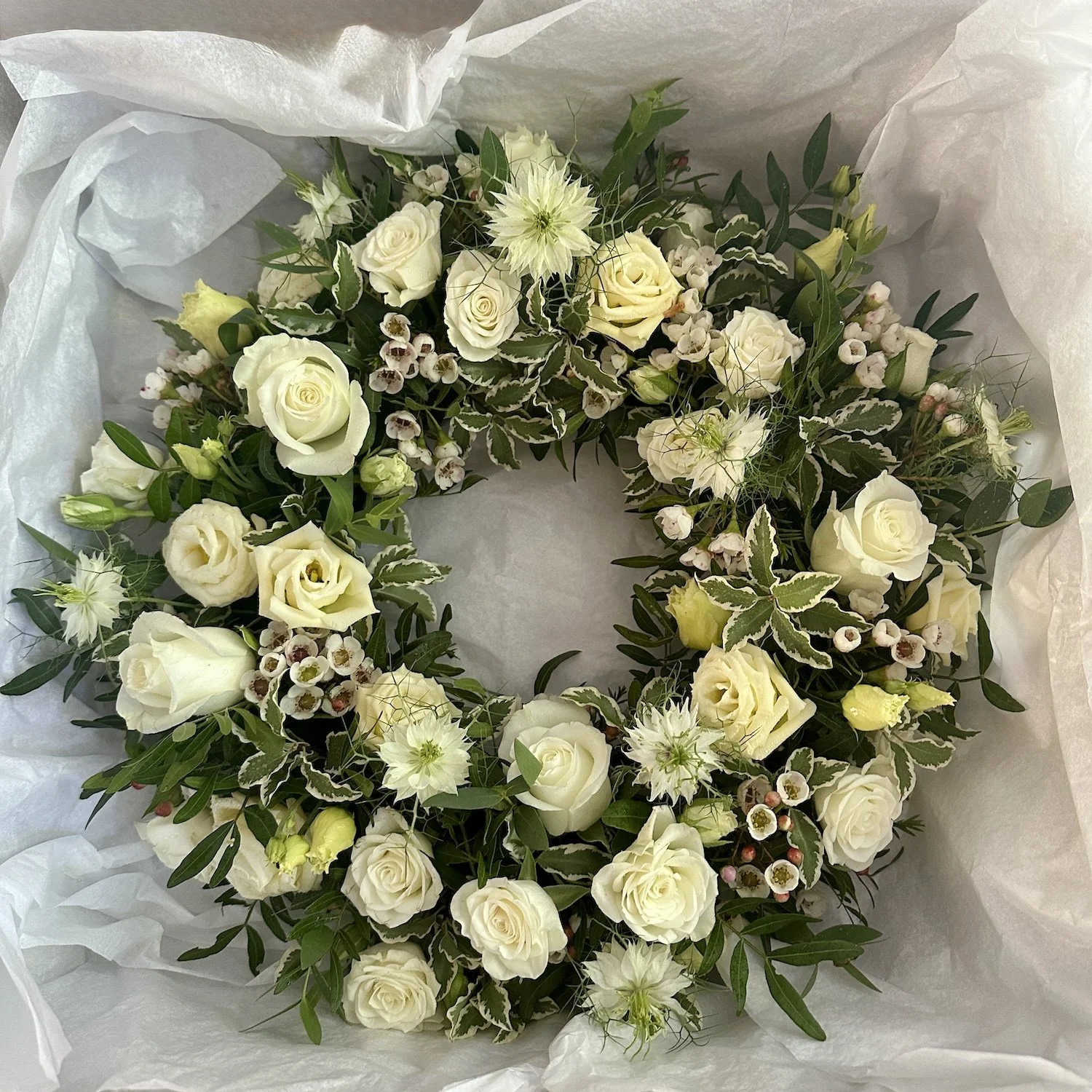White floral wreath with roses, lisianthus, and greenery, wrapped in white tissue paper.