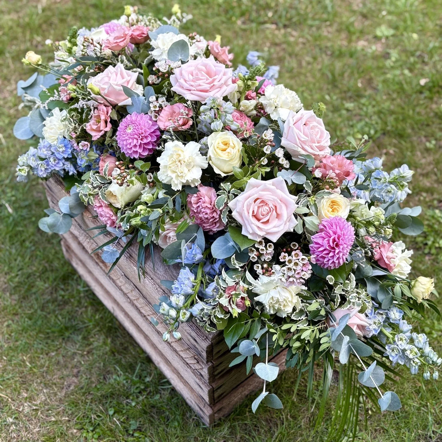 A floral casket spray arrangement with pink, white, and blue flowers on a wooden bench outdoors.