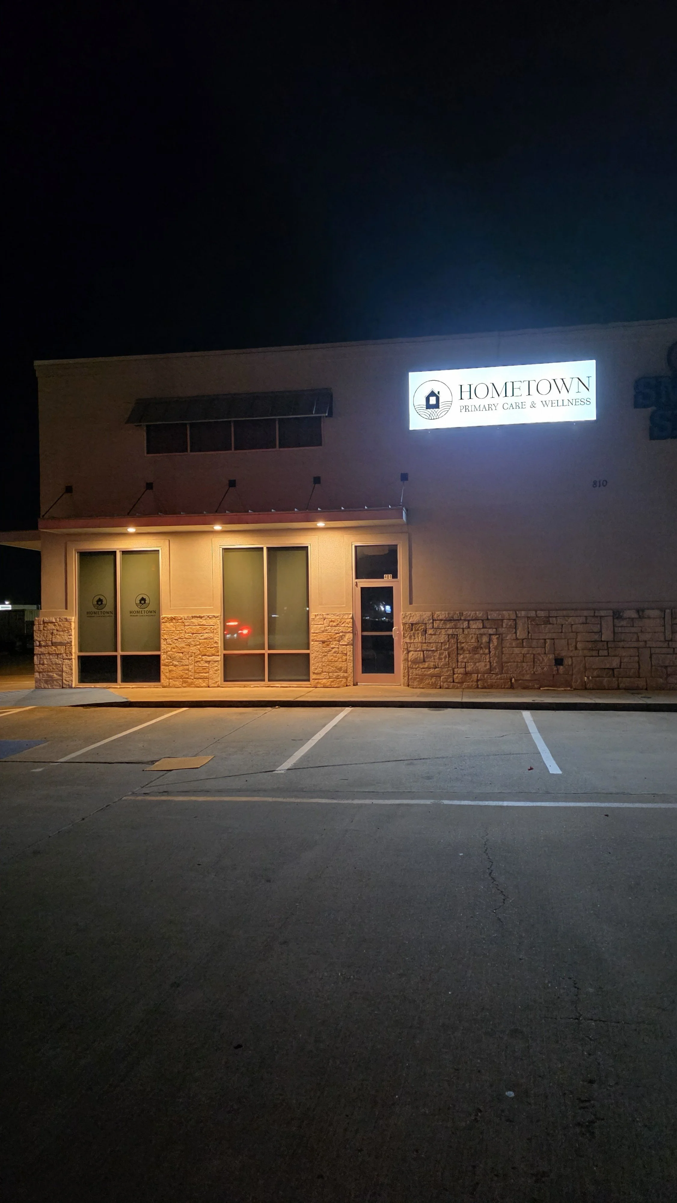 Nighttime exterior view of a building with a sign that reads 'Hometown Primary Care & Wellness' illuminated, with multiple parking spaces in front.