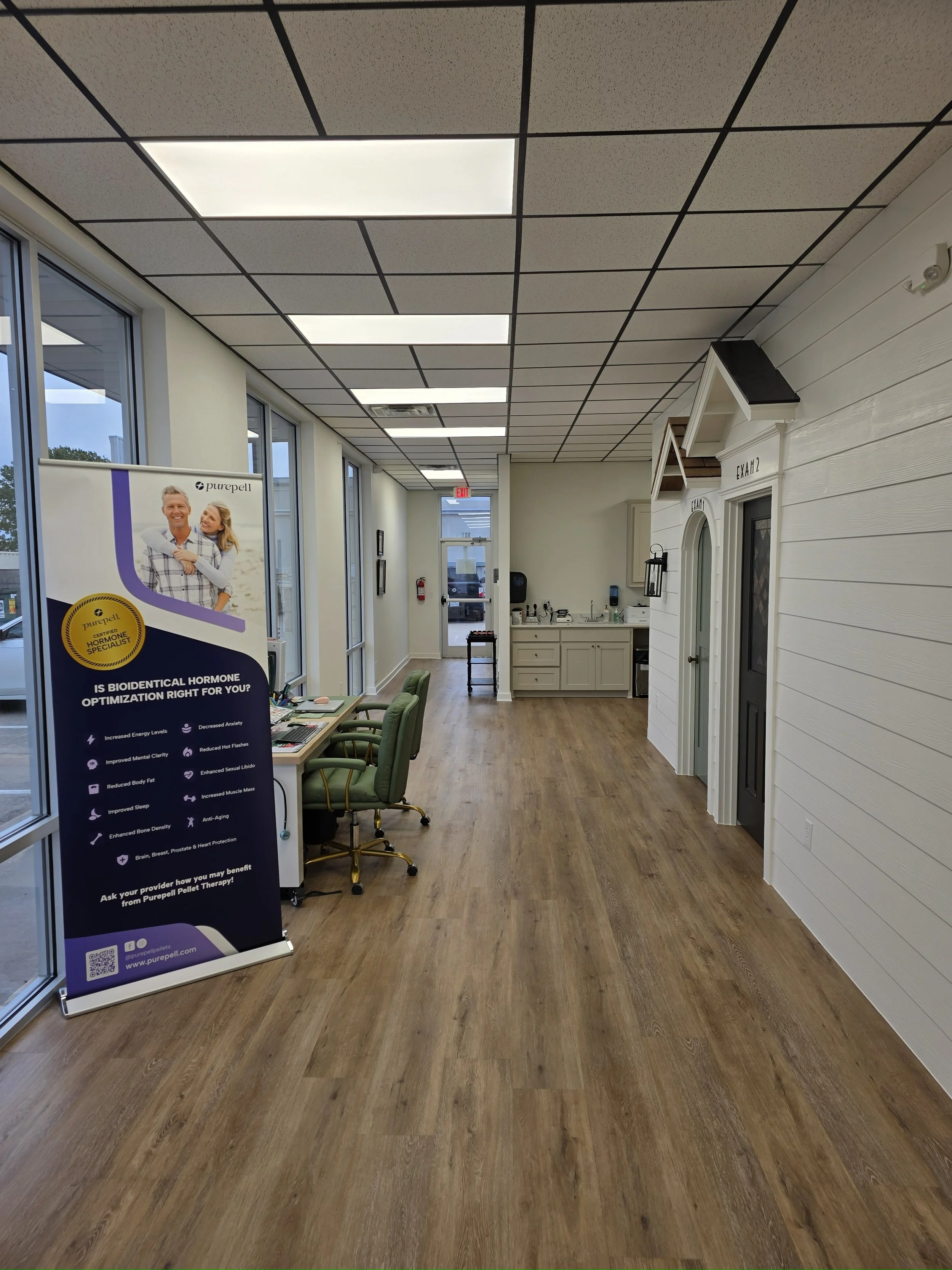 Interior of a medical or wellness office with a sign about bioidentical hormone optimization, chairs at a table, a kitchen sink area, and two exam rooms labeled 'Exam 1' and 'Exam 2'.