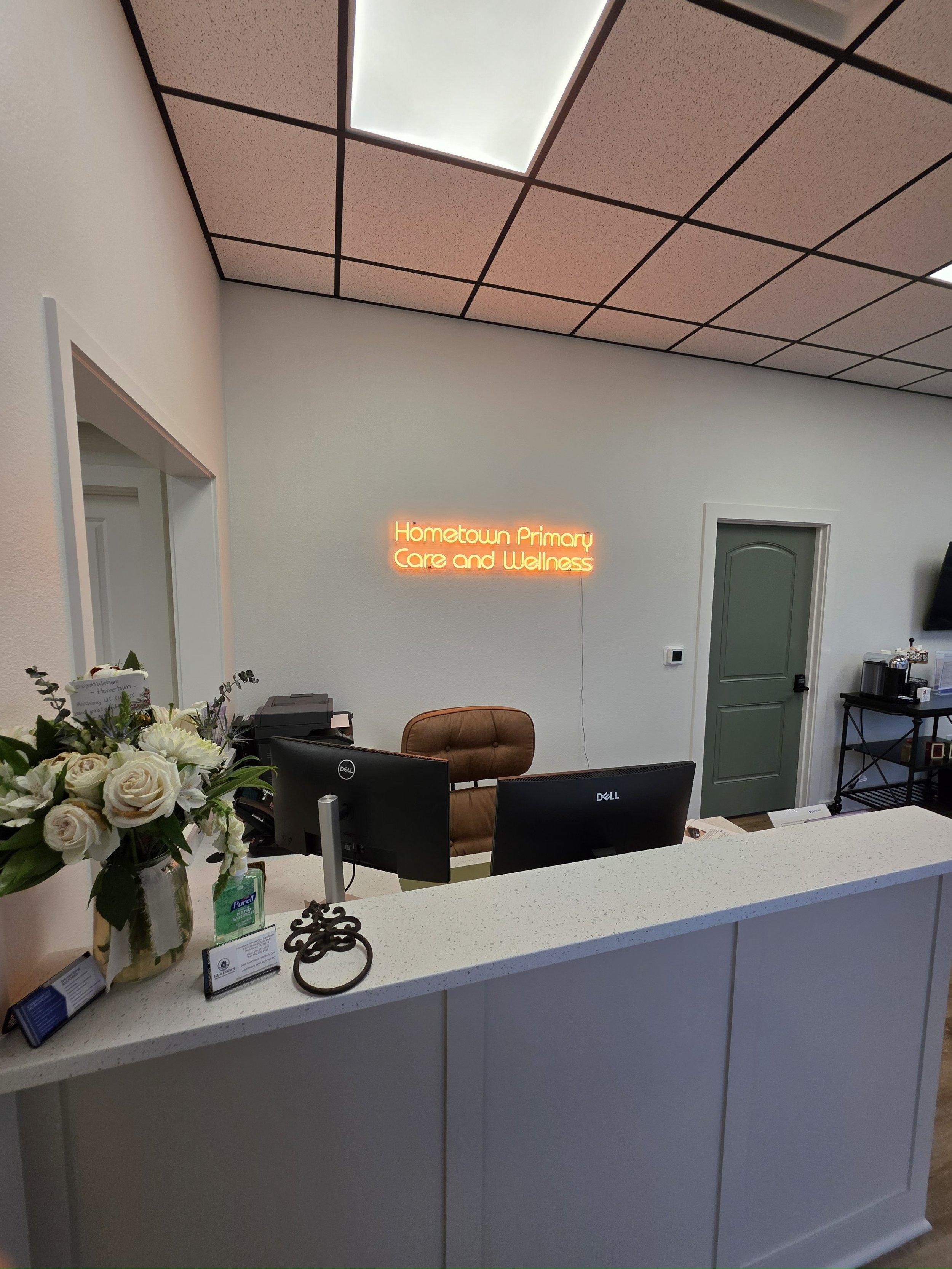 Reception area of Hometown Primary Care and Wellness with a white countertop, flowers, a sign, two computer monitors, and a brown chair behind the desk. Neon sign on the wall reads 'Hometown Primary Care and Wellness'.