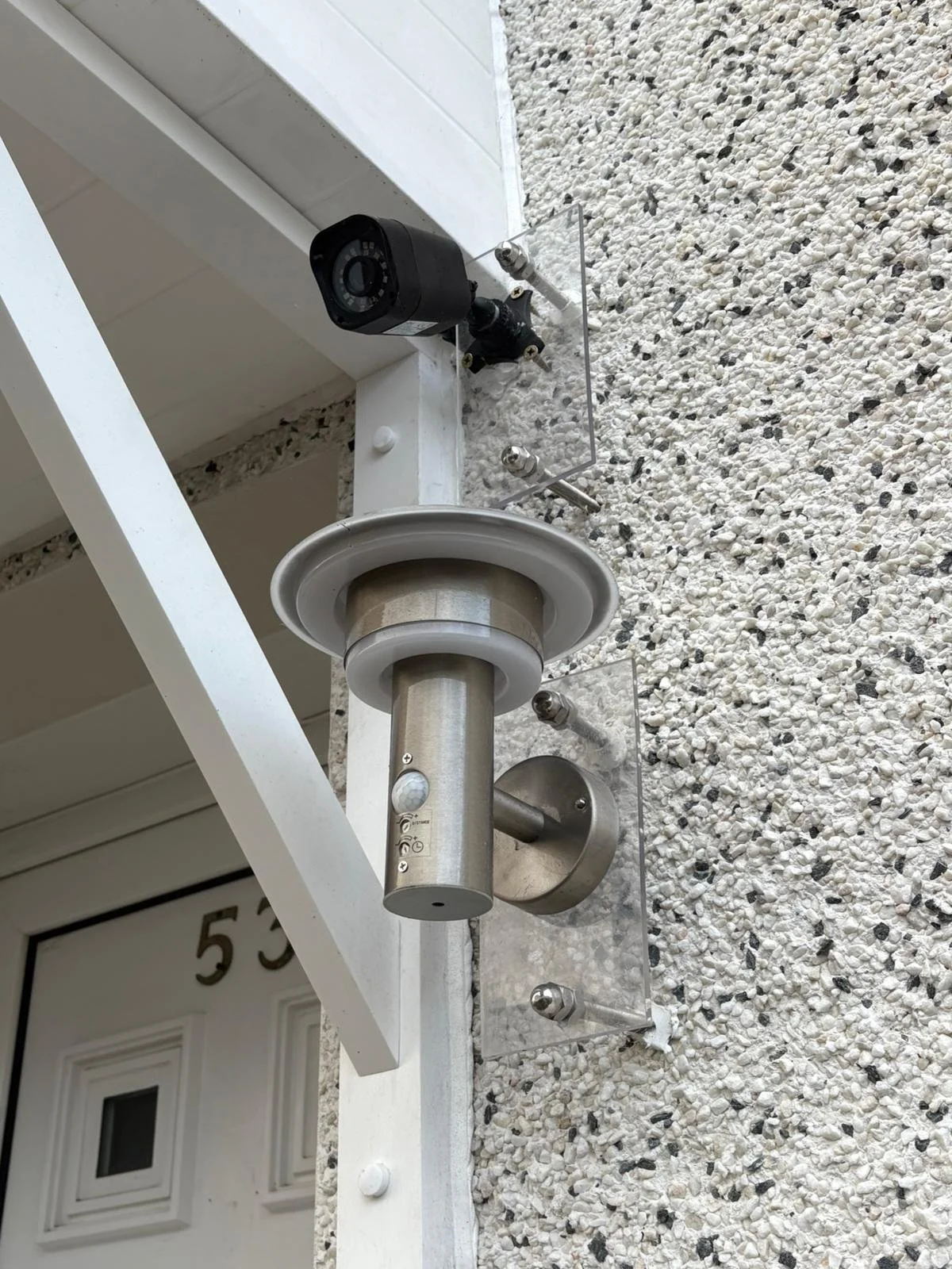 Close-up of metallic doorbell and security camera on a textured white exterior wall.