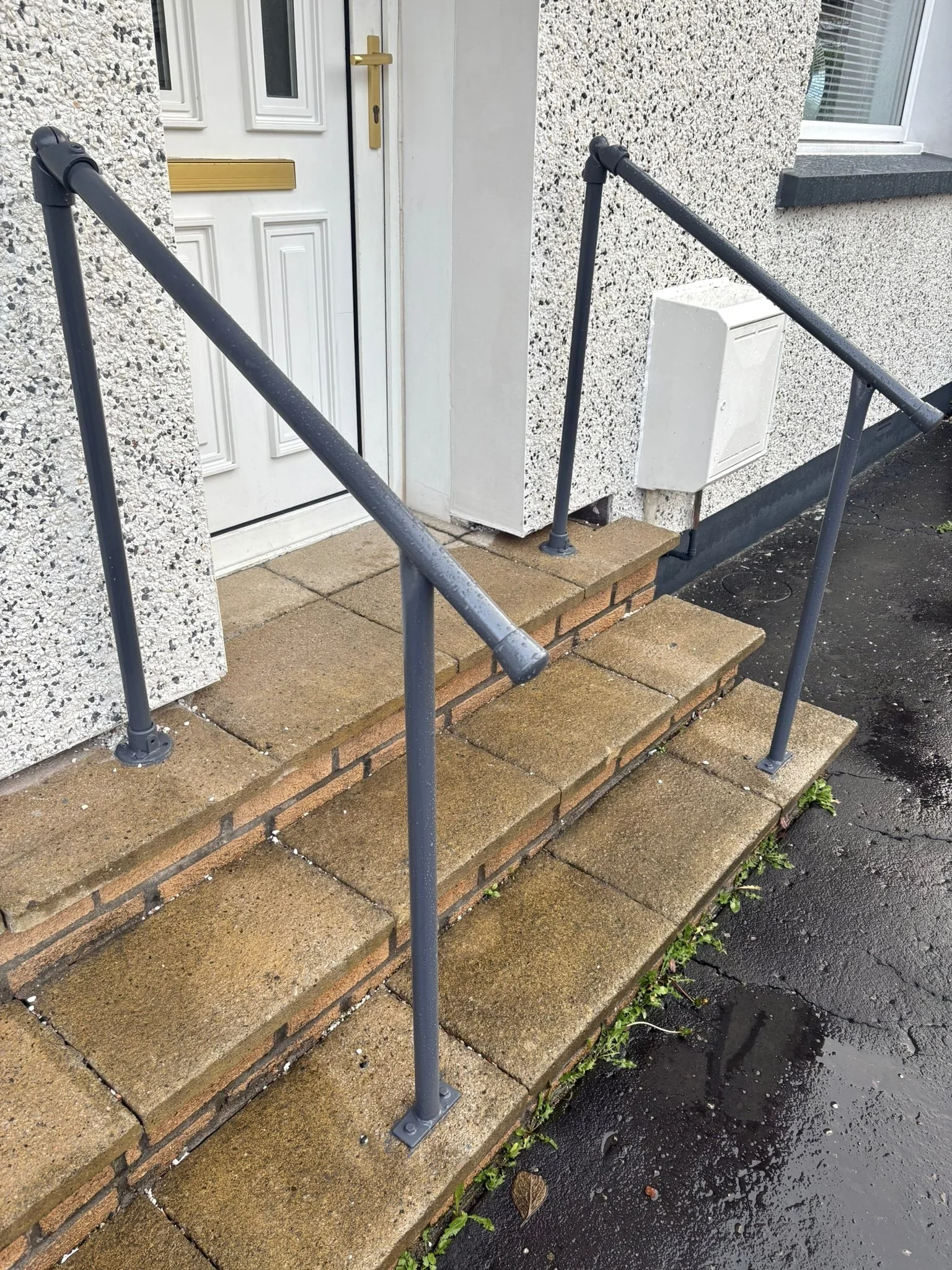 Brick steps leading to a white front door with a gold handle, black railing on both sides, and a small white utility box attached to the house wall, with wet black pavement in the foreground.