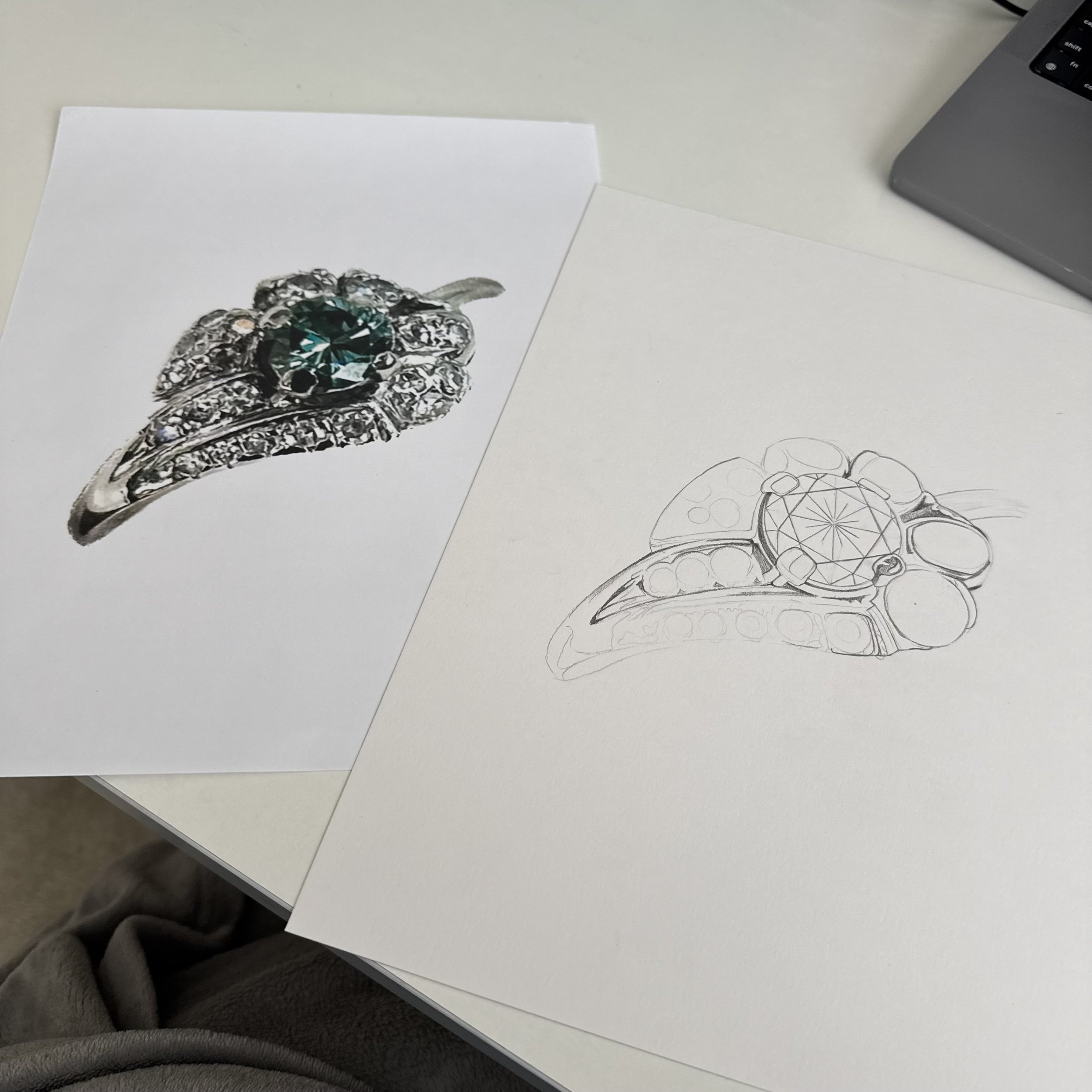 In progress photo of a black-and-white custom drawing for an engagement ring with a large central gemstone and intricate metalwork, next to the pencil sketch is a color photo reference on the artist's desk.