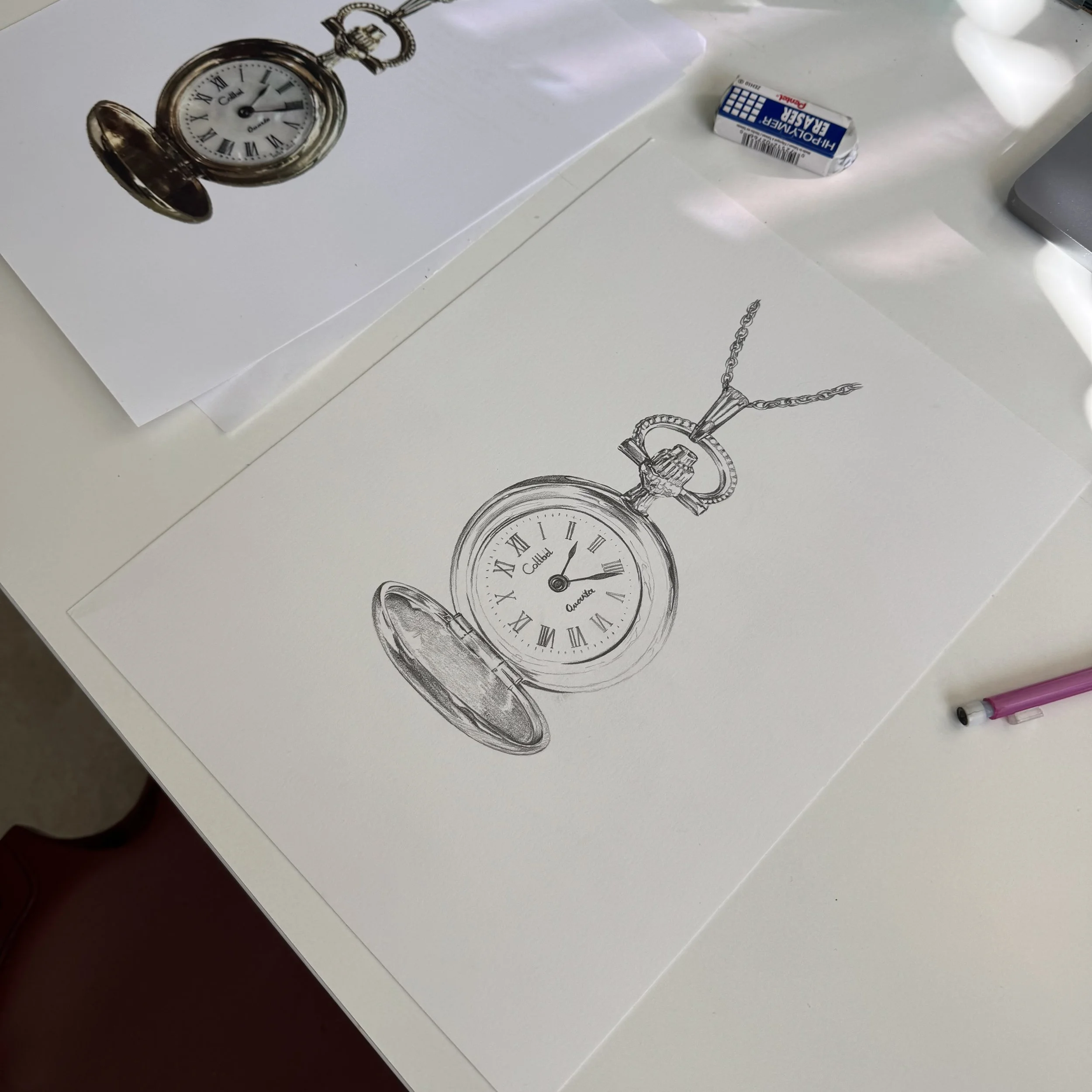 An artist's table with a printed image of a pocket watch, alongside a working sketch of the same pocket watch showing Roman numerals. An eraser and mechanical pencil are on the desk in the background.