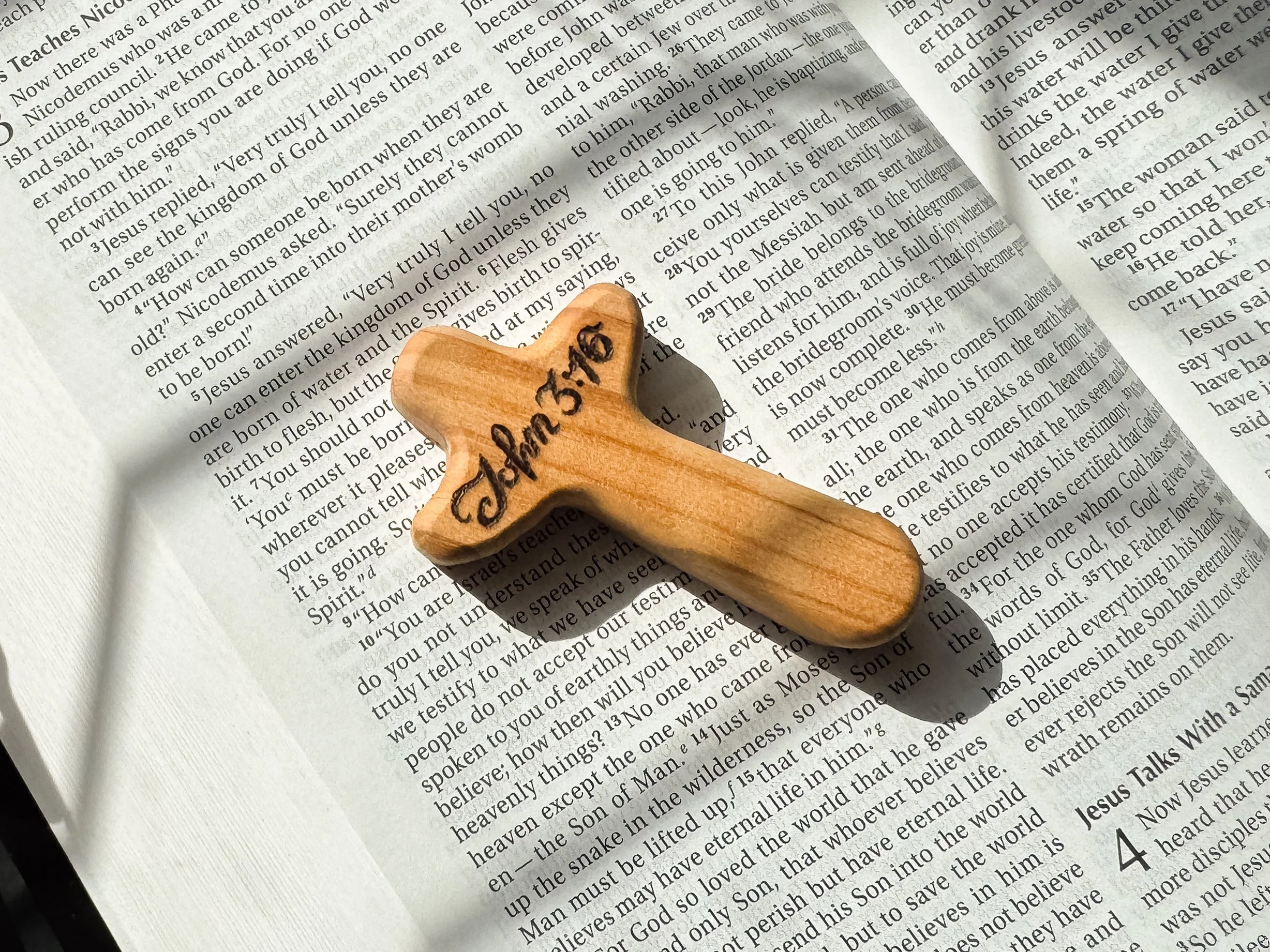 A wooden pocket cross with hand lettering 'John 3:16' engraved on to it. The gift sits over an open Bible and is illuminated by sunlight.
