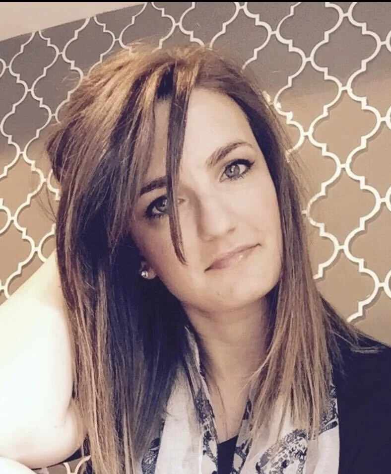 A young woman with light brown hair and blue eyes posing in front of a decorative patterned wall.